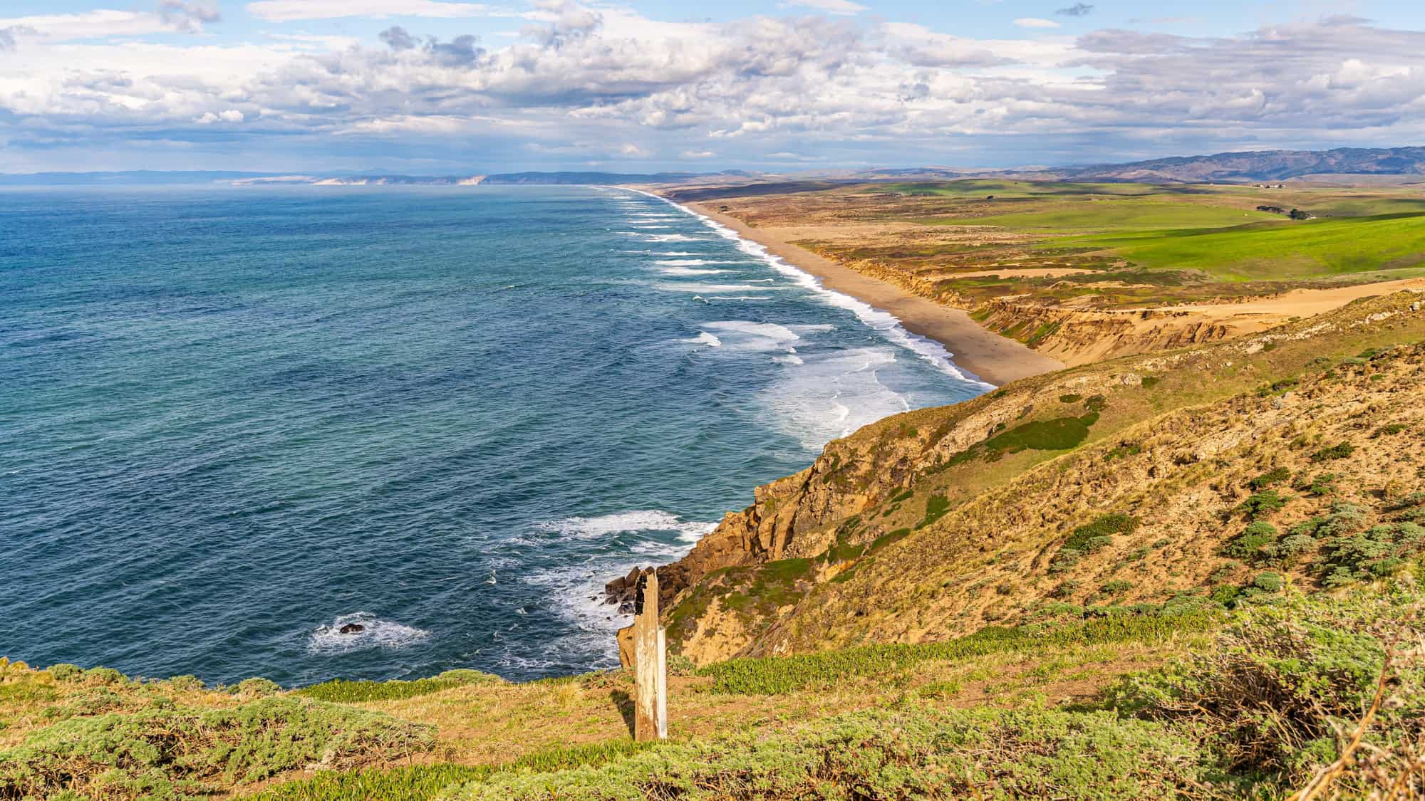 An aerial view of Point Reyes National Seashore shows endless waves meeting a golden shoreline under a partly cloudy sky, with green pastures sprawling inland.
