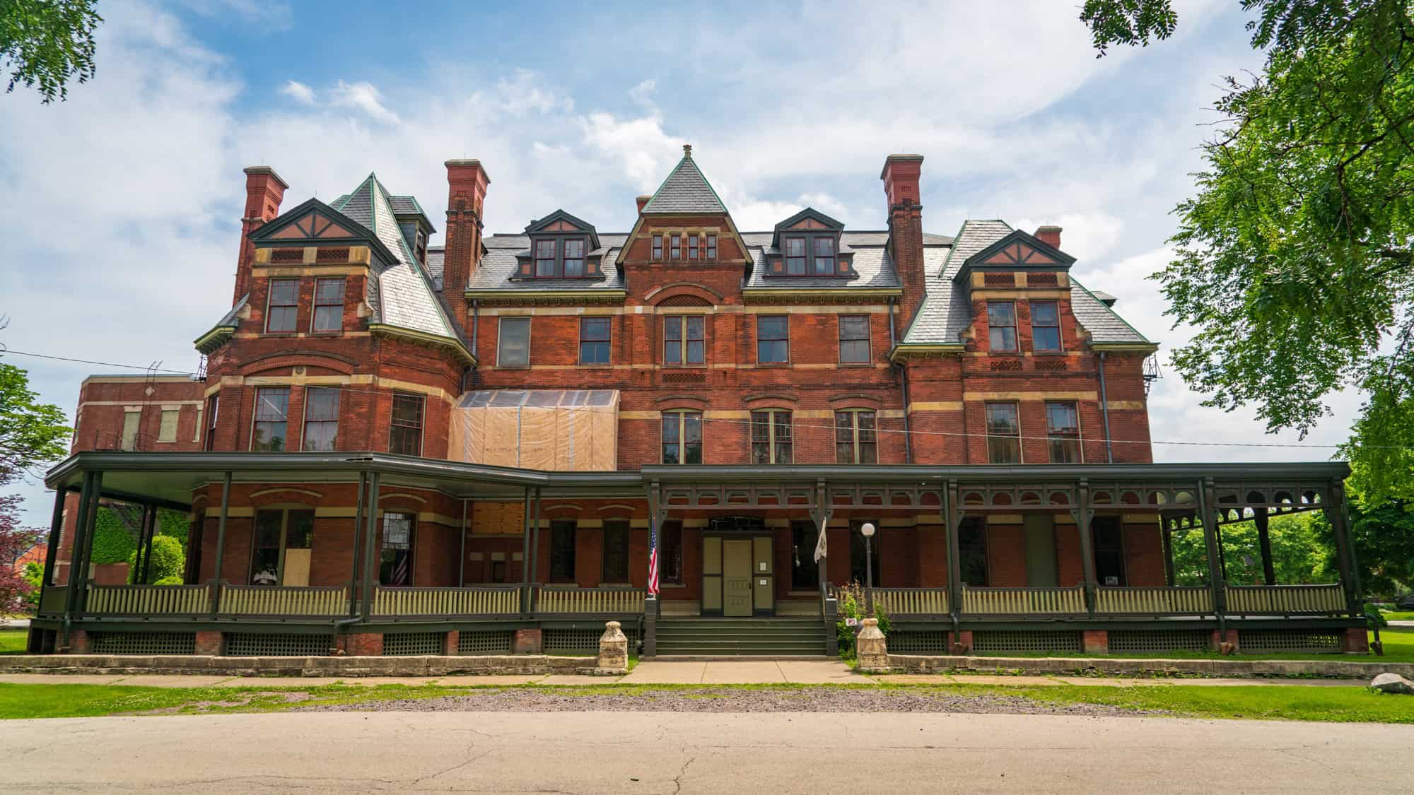 A grand, red-brick Victorian building with green-trimmed gables and tall chimneys sits under a bright sky, partially under renovation with boarded windows and scaffolding on one side.