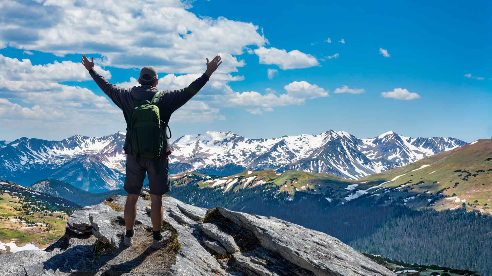 A hiker with raised arms stands triumphantly on a rocky outcrop overlooking snow-speckled peaks and alpine meadows in Rocky Mountain National Park.