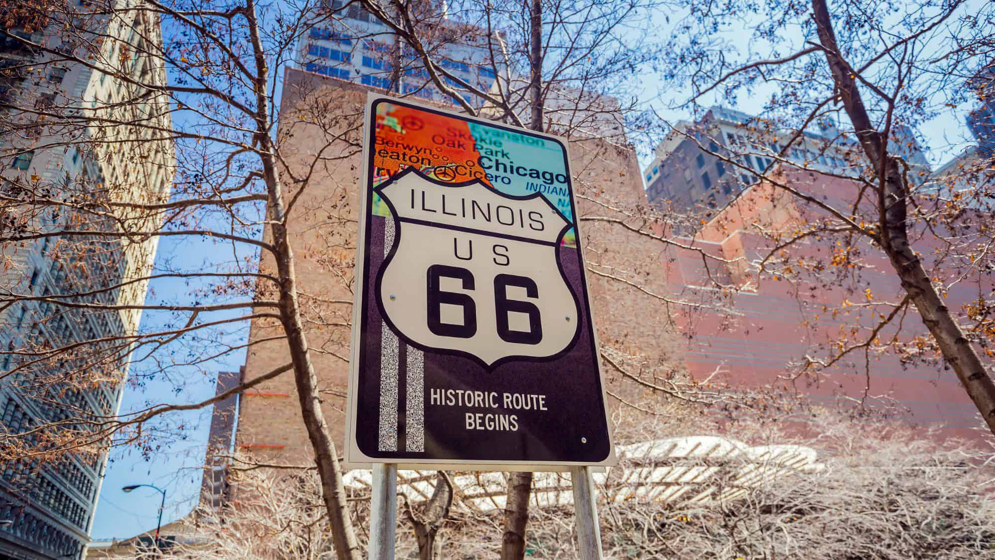 A street-level view of a sign that reads “Illinois US 66 Historic Route Begins” set against tall downtown Chicago buildings and bare winter trees.