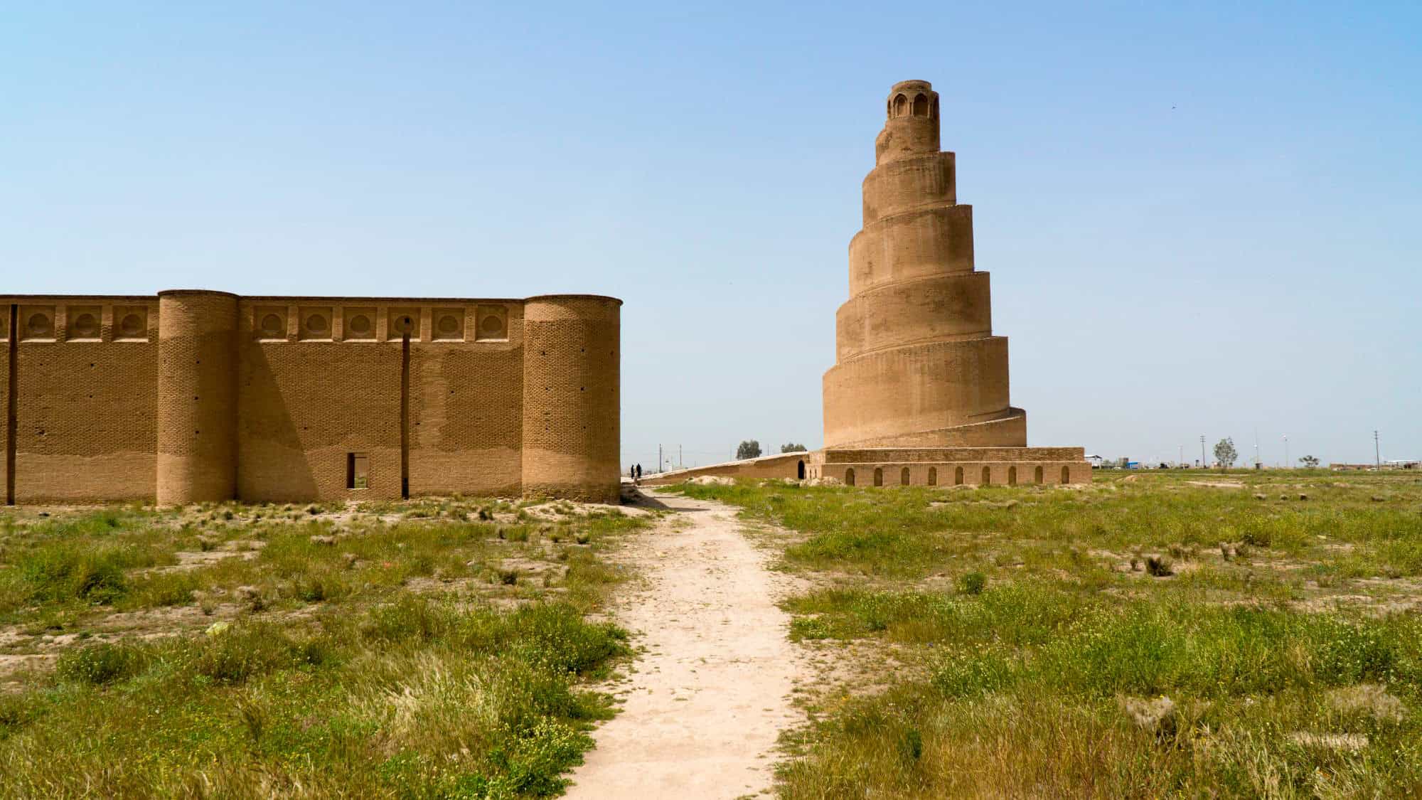 The ancient spiral minaret of the Great Mosque of Samarra rises dramatically from the desert landscape in Iraq, flanked by weathered brick walls.