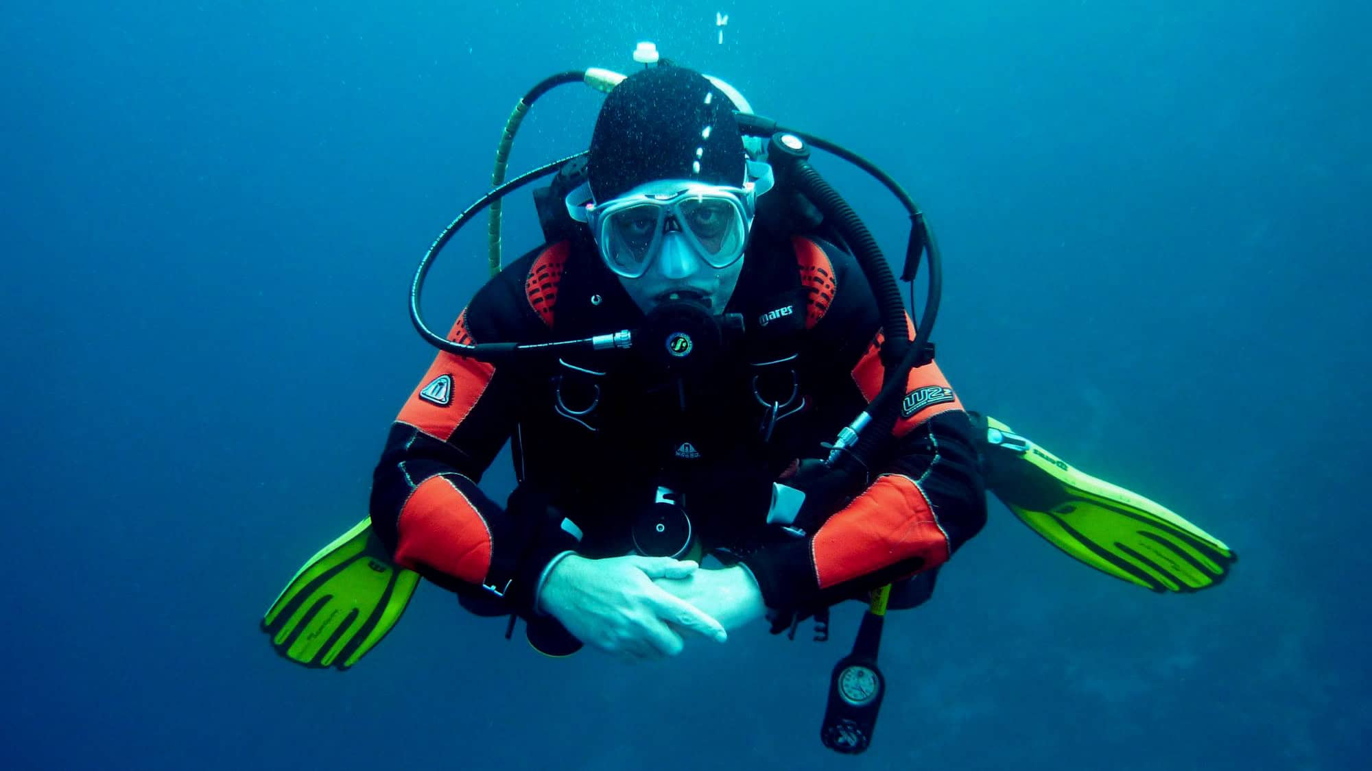 A diver in a red and black wetsuit with yellow fins floats underwater in a murky blue environment, facing the camera with arms crossed and surrounded by diving gear.