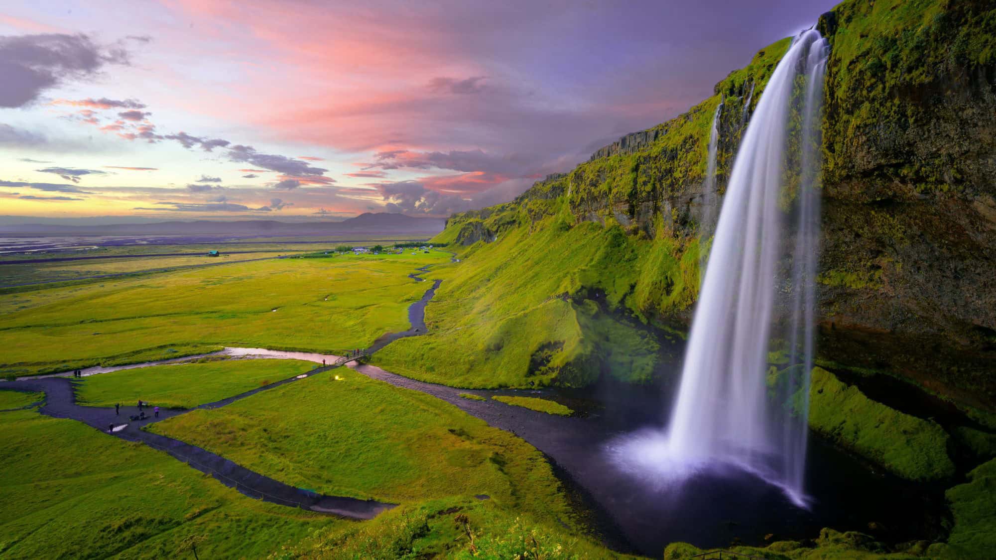 A narrow, tall waterfall flows off a mossy cliff onto the green Icelandic plains, with a pastel-hued sunset sky stretching into the distance.