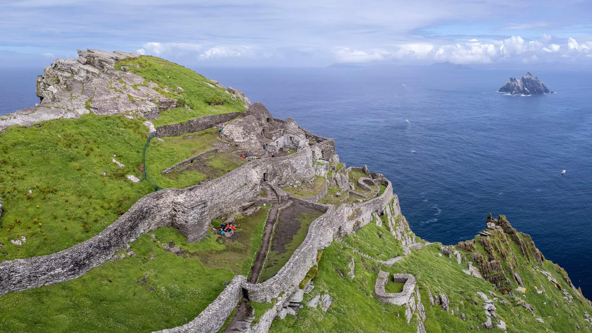 Ancient stone structures of a monastery scattered across the grassy, rocky slopes of Skellig Michael island, with the Atlantic Ocean in the background.