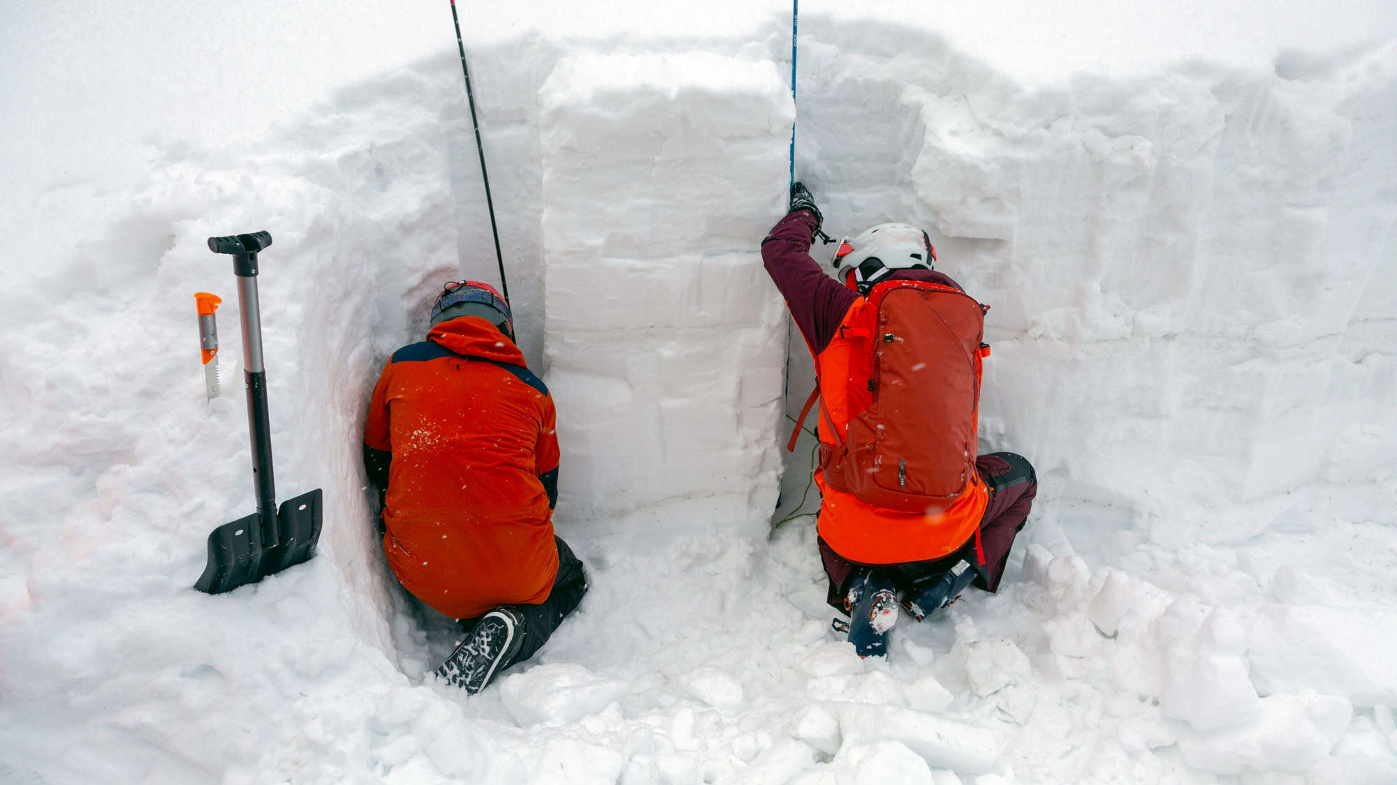 Two people in bright jackets dig into a deep snow trench for avalanche testing, with gear like shovels and poles around them in a thick, snowy landscape.