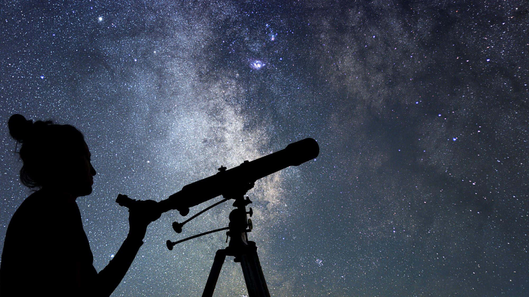 Silhouette of a person looking through a telescope under a vast, star-filled night sky, with the Milky Way glowing vividly overhead.