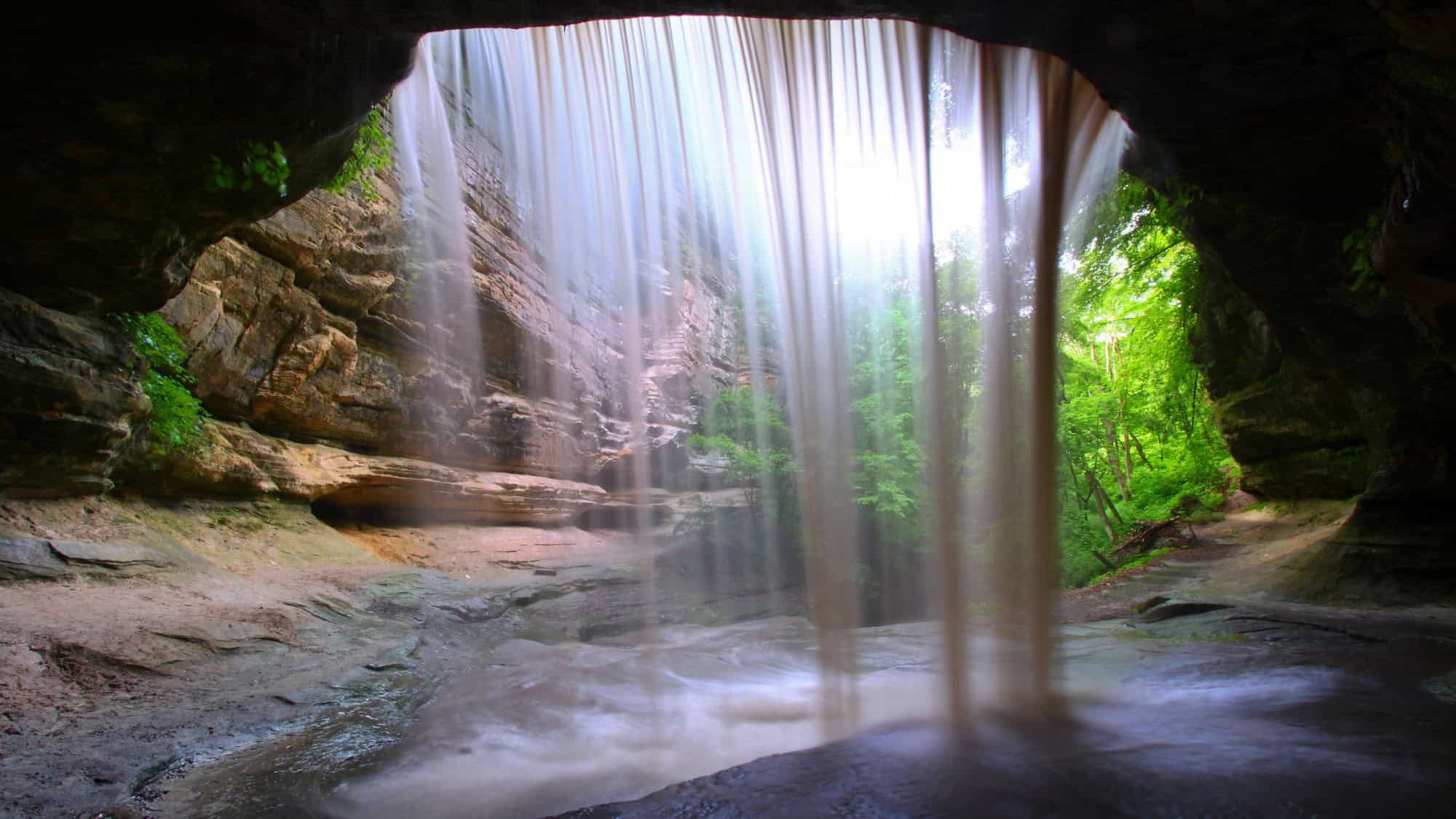 A dramatic view from behind a silky waterfall as it streams over the edge of a rock ledge, surrounded by mossy cliffs and lush green forest.


