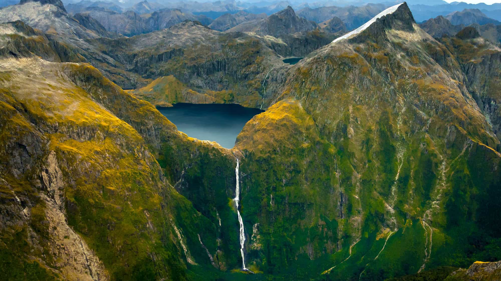 Remote alpine scenery with a slender, tall waterfall descending from a hidden lake nestled among steep, rugged mountains.