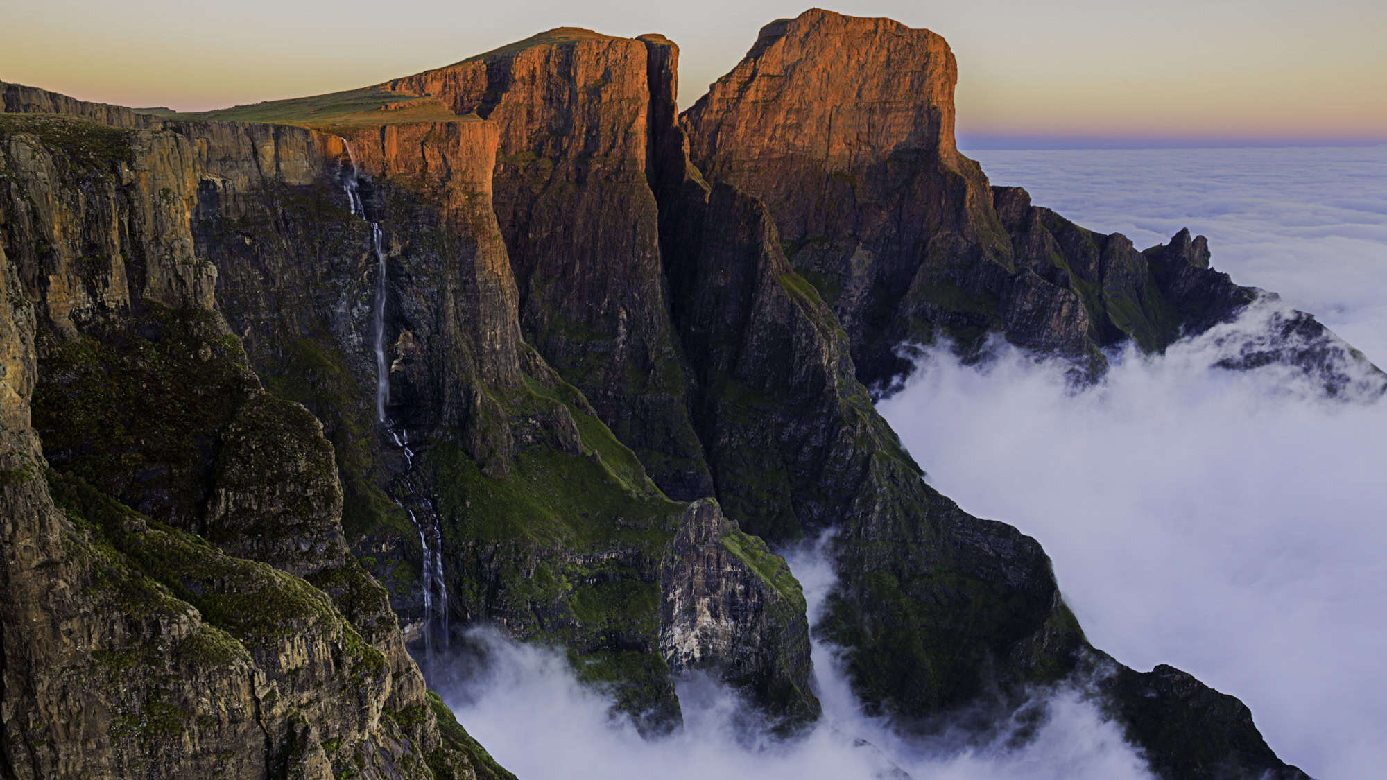 Towering cliffs with Tugela Falls streaming down the rock face, partially hidden by low clouds blanketing the valley below.