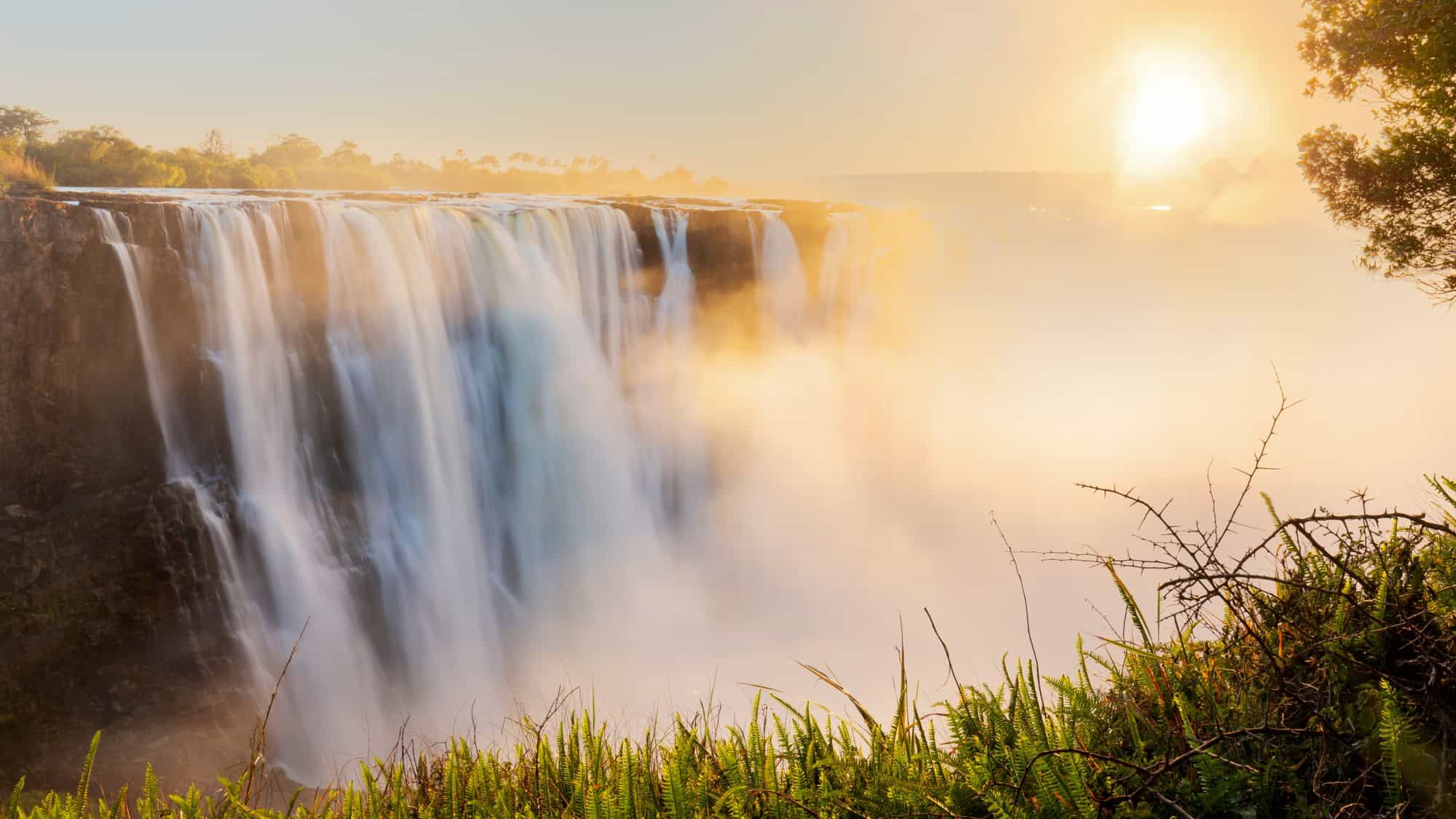 A majestic view of Victoria Falls cascading over a wide cliff edge, with golden sunlight beaming through the mist and lush greenery in the foreground.