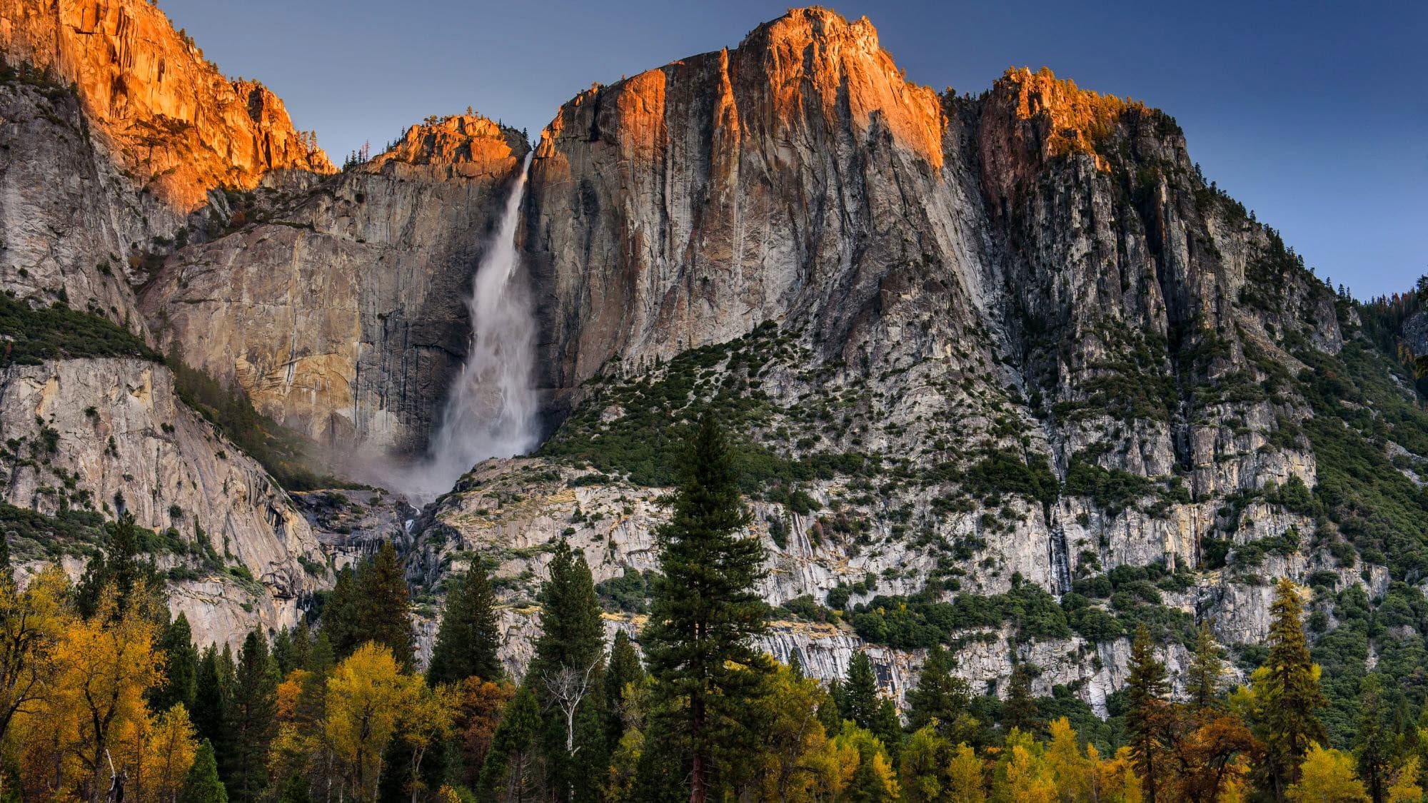 A powerful column of water falls from the granite cliffs of Yosemite National Park, framed by golden autumn trees below. The light of sunset warms the towering rock faces, highlighting the grandeur of this iconic U.S. landscape.