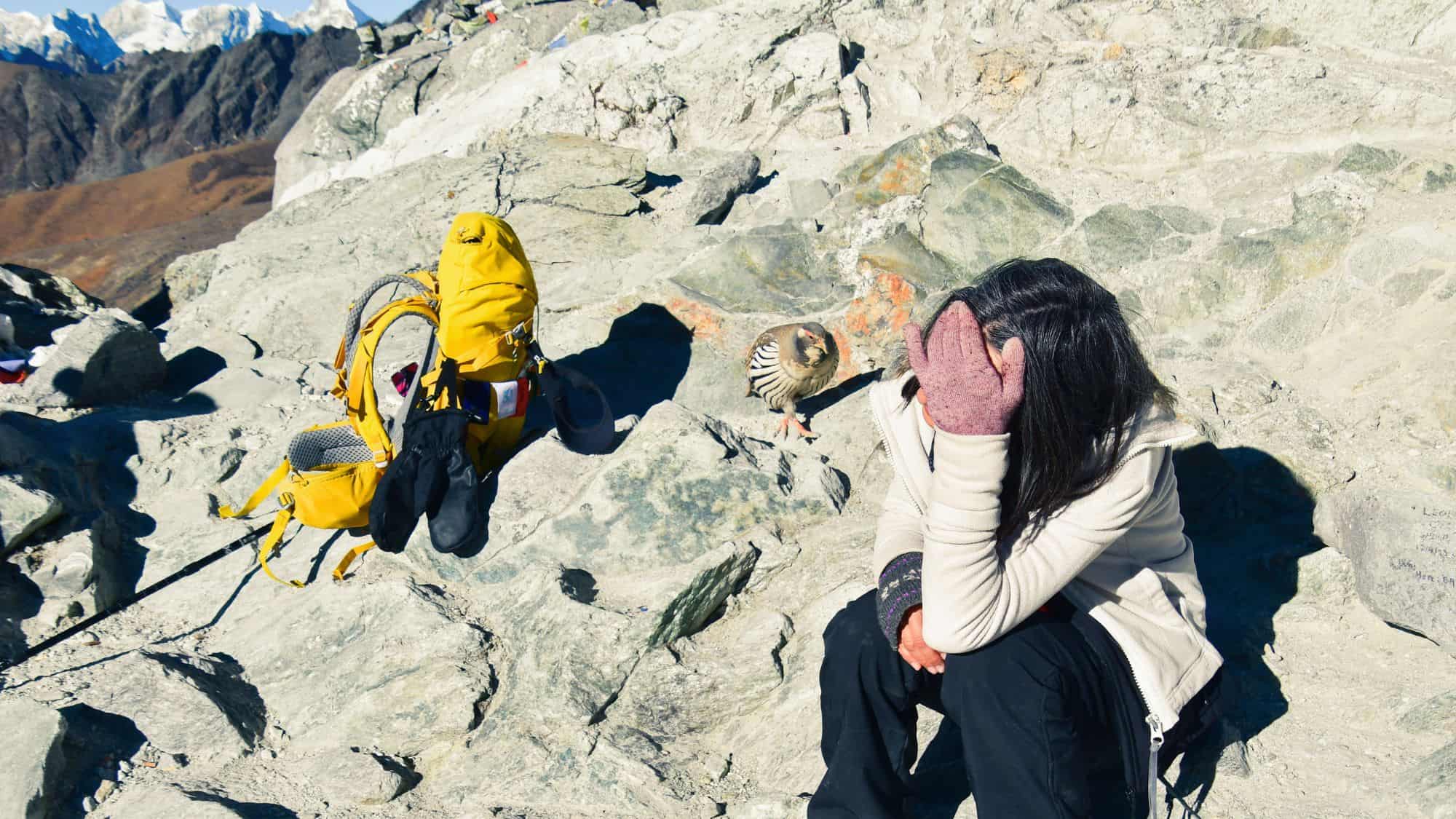 A hiker sits on a rocky mountain ledge with her head in her hand, appearing fatigued or unwell, possibly from altitude sickness, while a yellow backpack and trekking poles rest nearby and a chukar bird stands close to her.