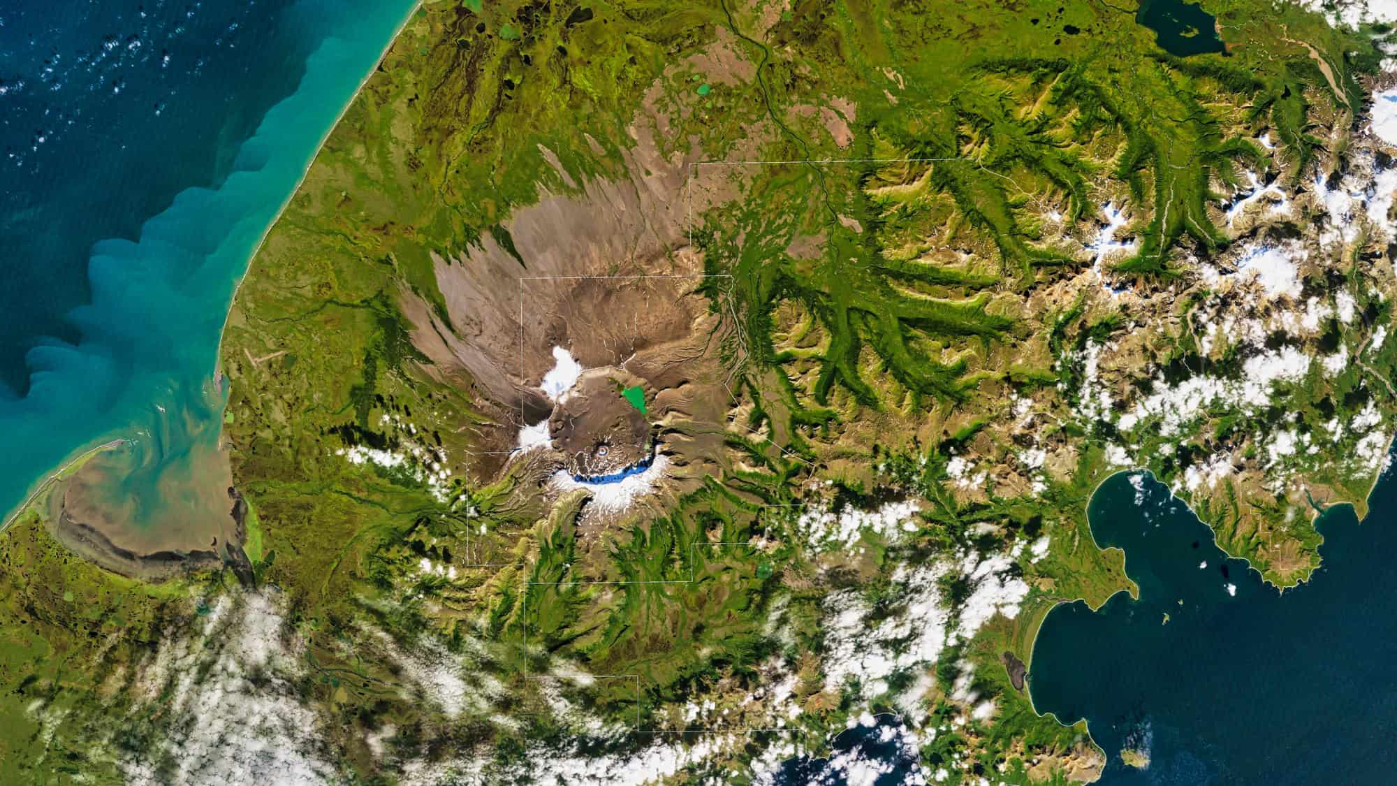 A satellite view of the rugged Aniakchak Caldera in Alaska, showing a massive volcanic crater ringed by green valleys, glacial remnants, and nearby coastal waters transitioning from turquoise to deep blue.