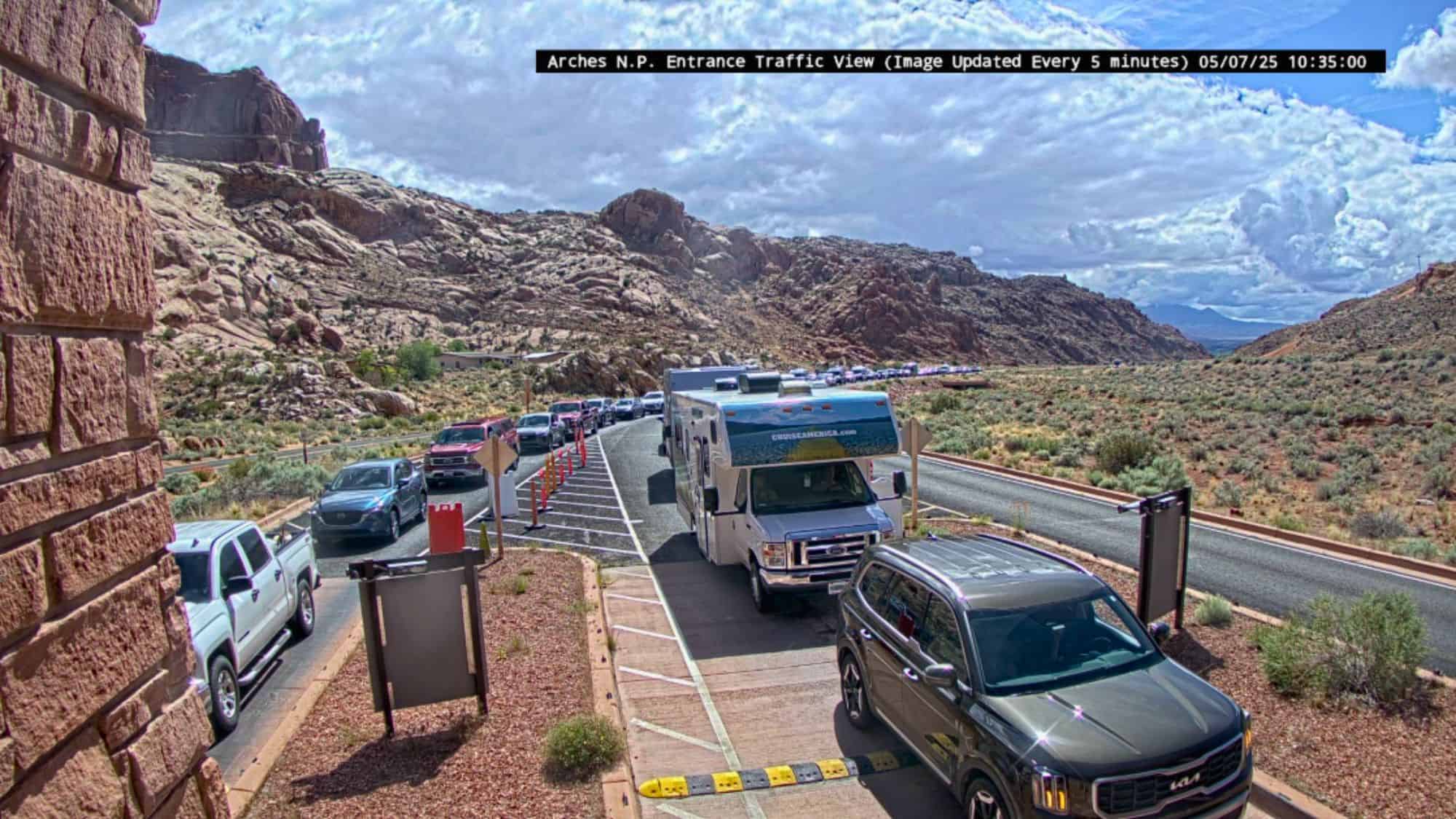 A long line of cars, RVs, and trucks waits at the Arches National Park entrance under a partly cloudy sky, surrounded by rugged red rock formations.