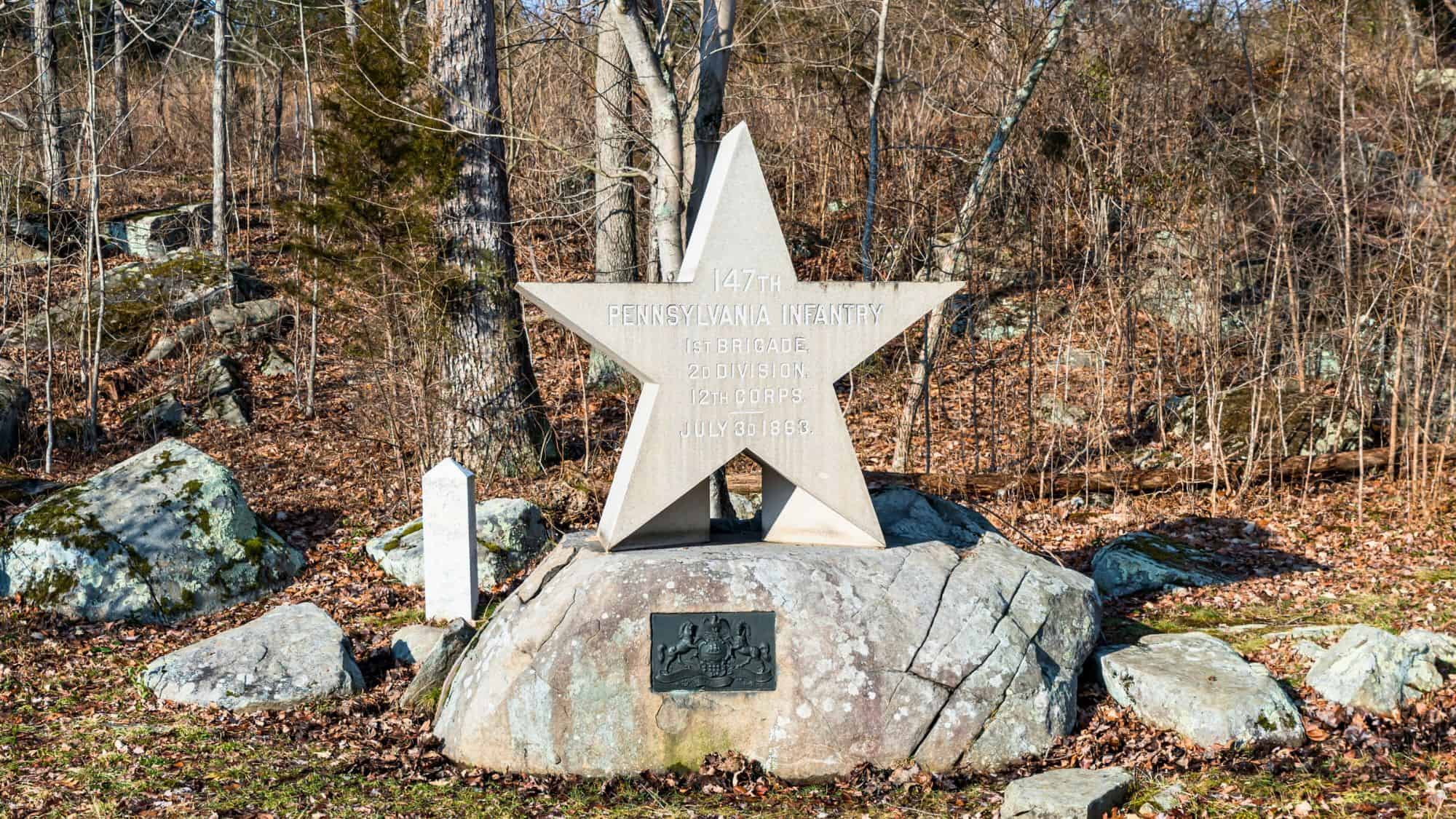 A stone star-shaped monument dedicated to the 147th Pennsylvania Infantry sits atop a large boulder in a wooded area, commemorating their role in the Battle of Gettysburg on July 3, 1863.