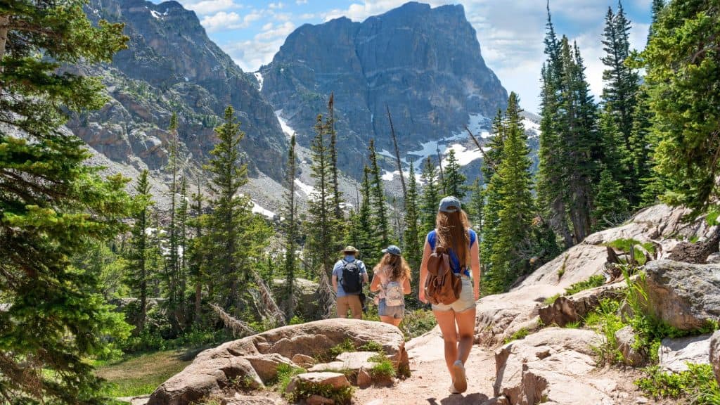 Group of hikers walking down a forested mountain trail in Estes Park, with towering rocky peaks and patches of snow in the background.