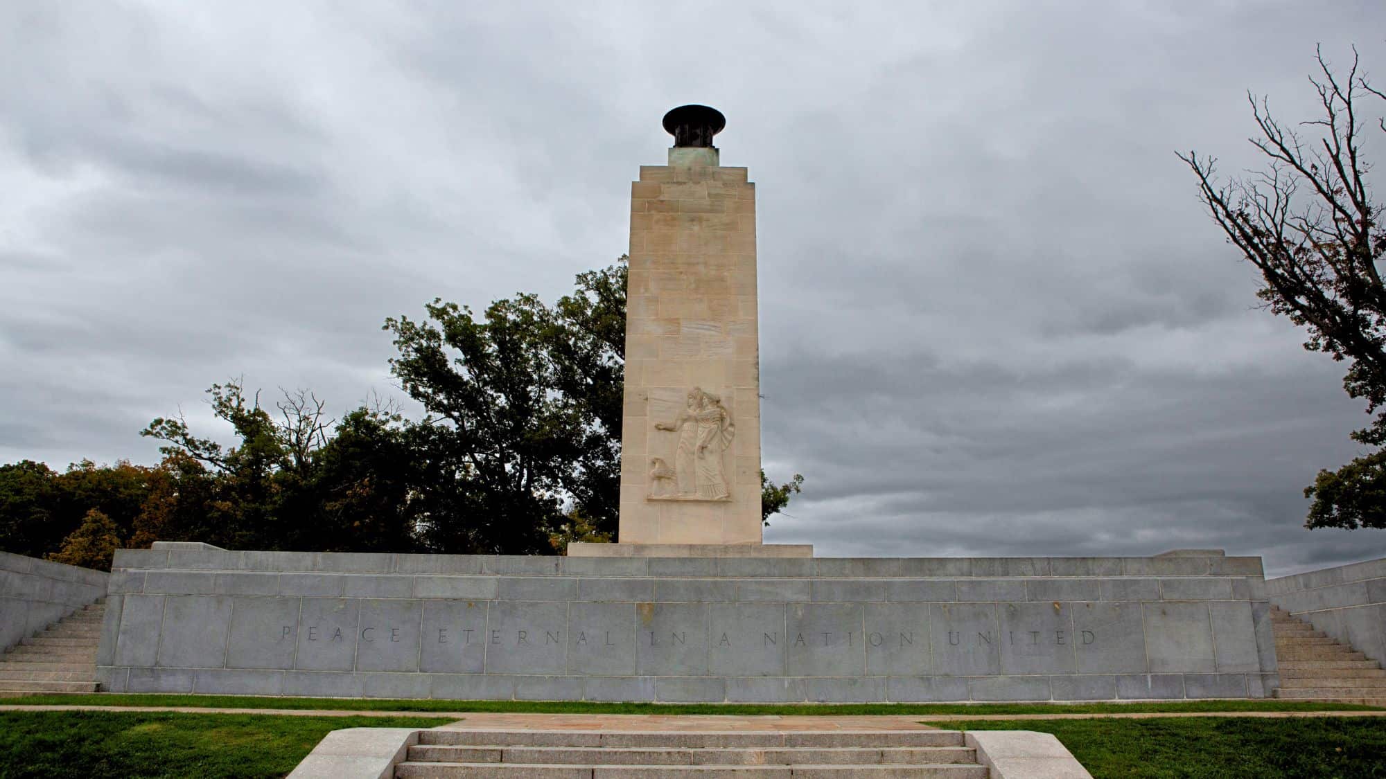A tall, stone monument with a bronze flame at the top stands against a cloudy sky; its base reads “Peace Eternal in a Nation United,” commemorating reconciliation after the Civil War.