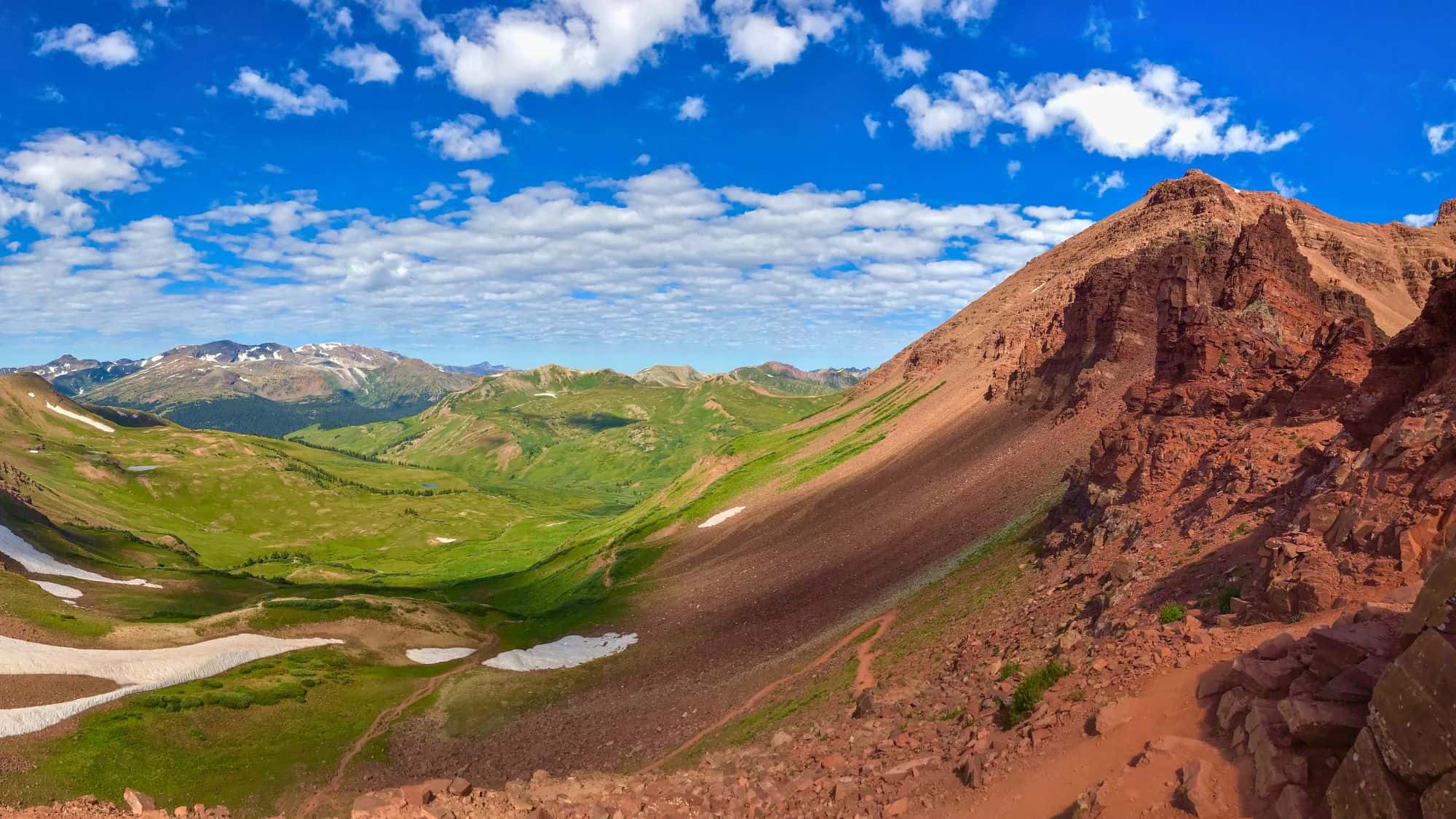 Expansive alpine landscape with green meadows, snow patches, and rugged red rock peaks under a bright blue sky filled with scattered clouds.