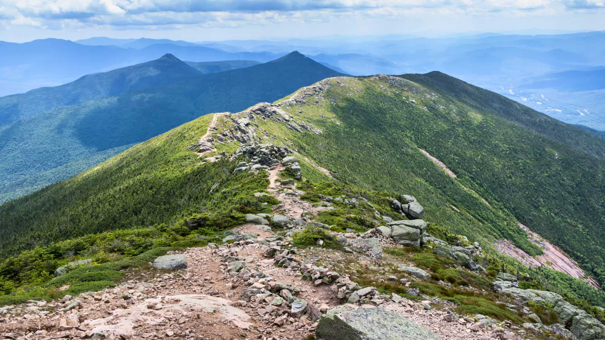 A rocky trail runs along a ridgeline with panoramic views of rolling, forested mountains stretching out under a hazy blue sky.