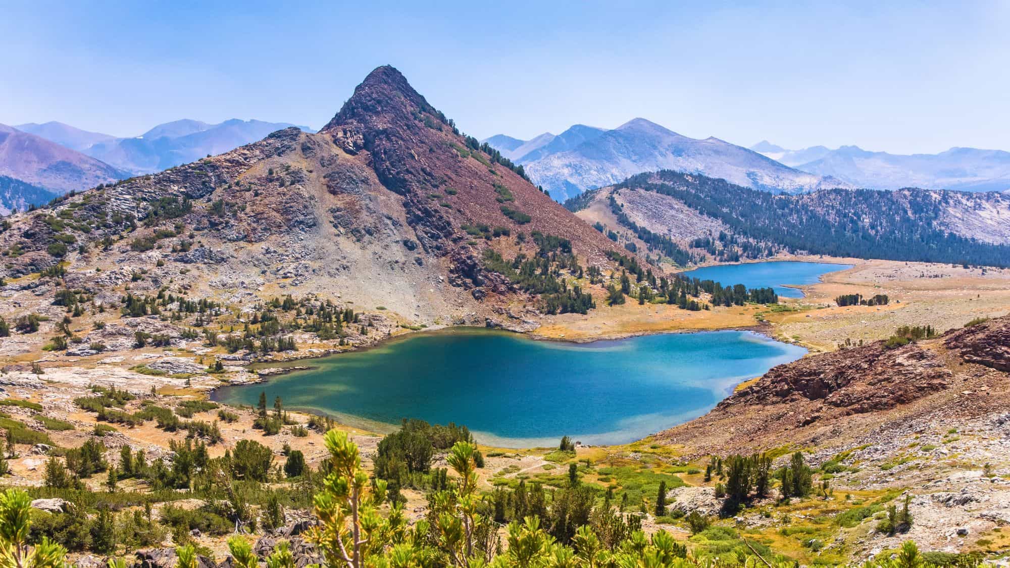 A panoramic view of Gaylor Lakes in Yosemite National Park shows two brilliant blue alpine lakes framed by rocky ridges and pine trees, with a jagged peak rising dramatically in the background.
