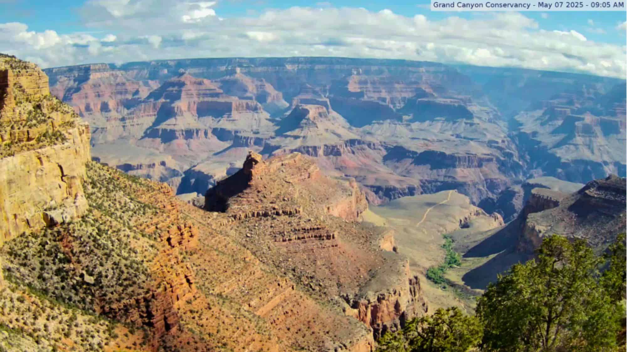 A sweeping view over the Grand Canyon reveals dramatic cliffs, layered rock formations, and a winding canyon floor under a sky dotted with clouds.