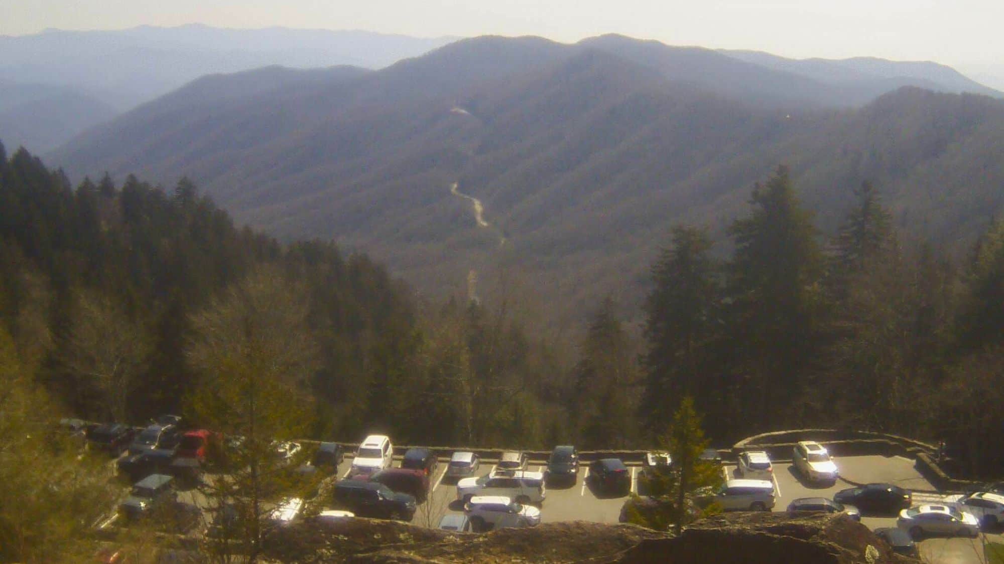 A parking lot packed with cars sits below a hazy mountain ridge, with winding roads cutting through the tree-covered Great Smoky Mountains.