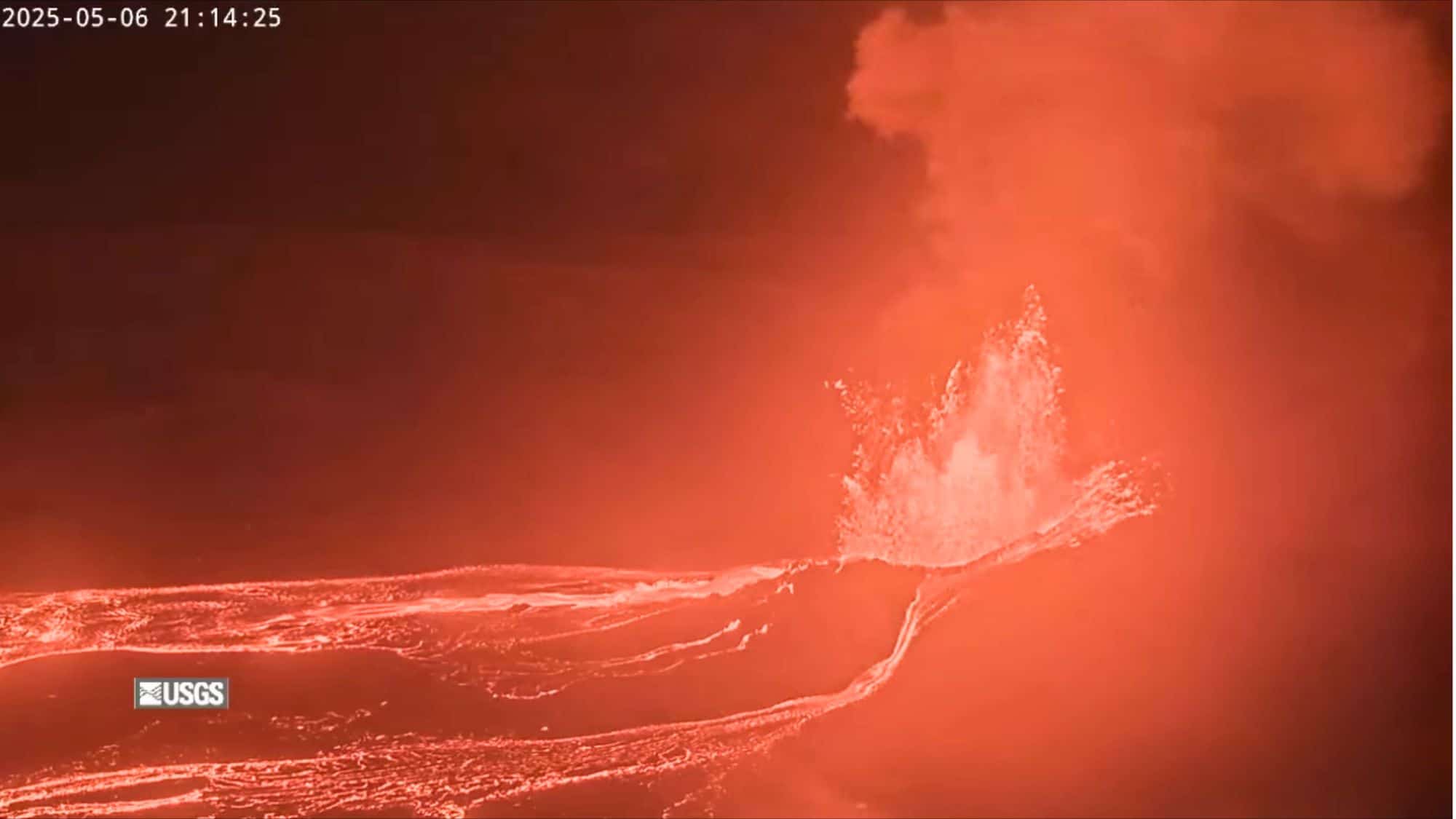 Bright orange lava erupts and flows across the dark landscape, with glowing sprays of molten rock under a nighttime sky, captured on a USGS webcam.