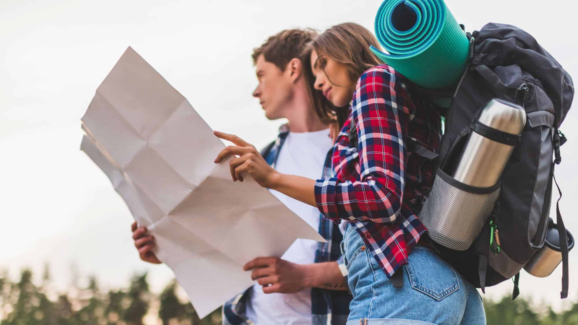 Two young hikers examining a paper map outdoors, dressed in casual hiking attire and carrying large backpacks with gear like mats and thermoses.