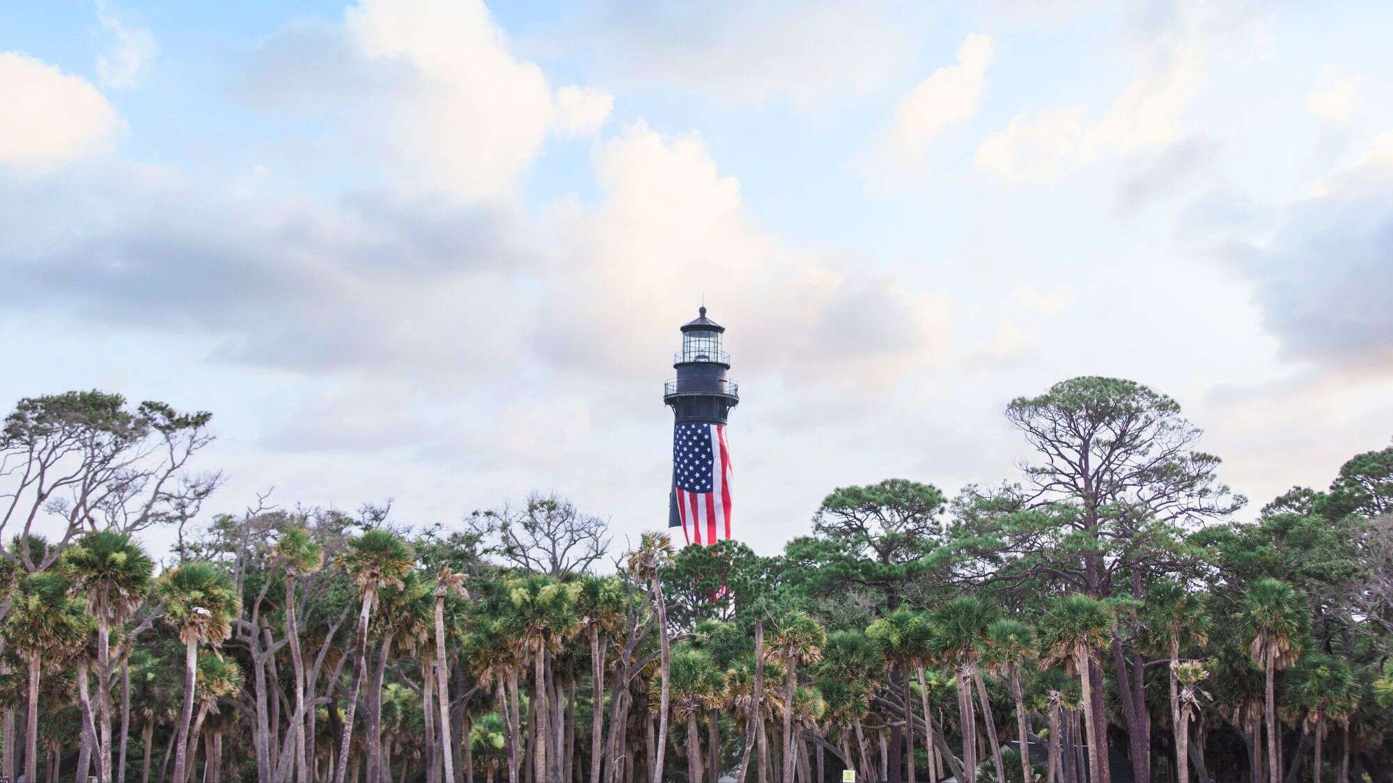 A historic black lighthouse rises above a dense forest of palm trees, with a large American flag draped down its front, under a sky of soft clouds.
