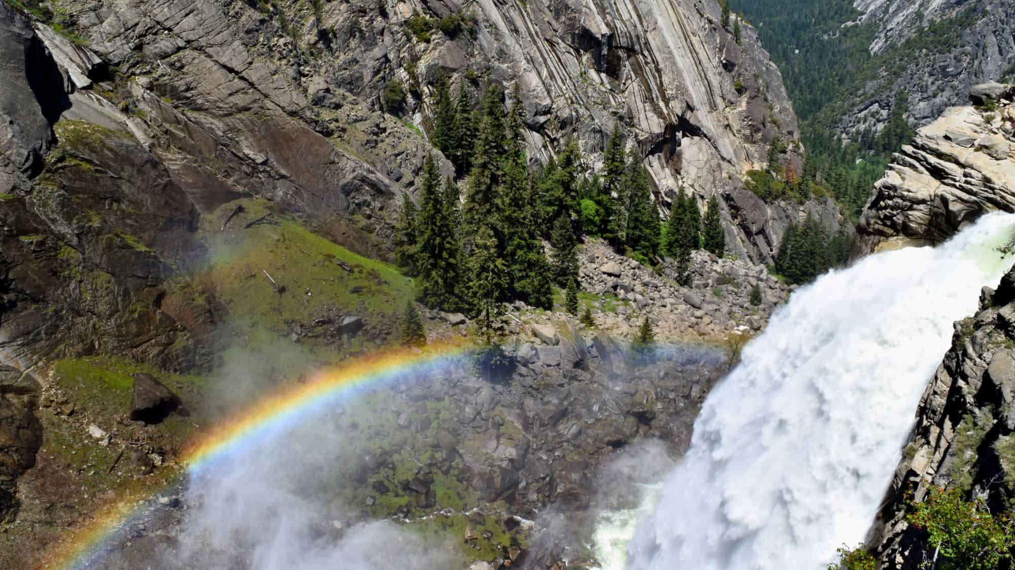 Water crashes over the edge of Illilouette Fall in Yosemite, with a vivid rainbow forming in the mist and a lush forested canyon sprawling below.