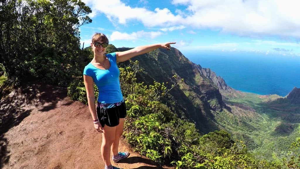 A smiling woman in athletic gear stands at a scenic overlook pointing toward the dramatic cliffs and turquoise ocean of the Nā Pali Coast.