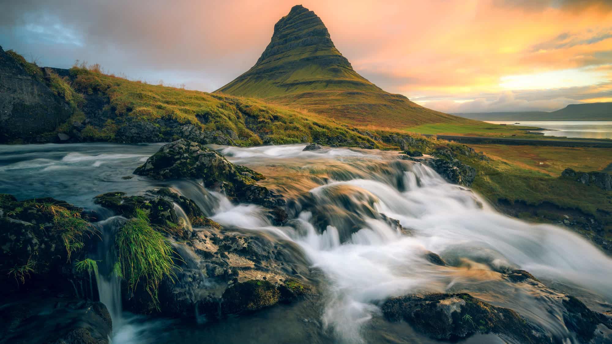 Kirkjufell Mountain rises with its iconic cone shape in the background, framed by cascading waterfalls and a glowing sunset sky. The water in the foreground flows over dark rocks, adding depth and motion to this serene Icelandic landscape.