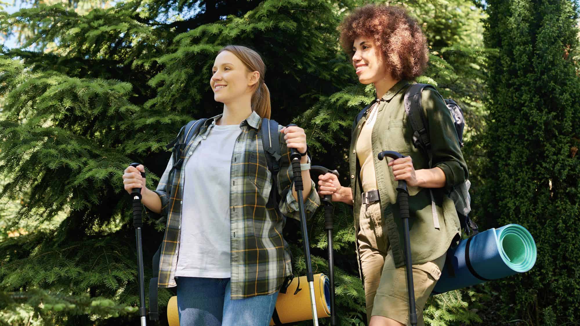 Two women hikers smiling in a forest, each wearing layered clothing and carrying rolled sleeping pads and trekking poles.