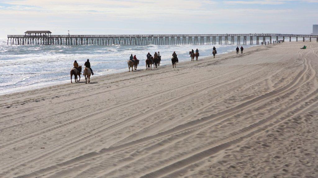 A line of horseback riders makes its way along the sandy shore with the Myrtle Beach pier stretching out into the ocean nearby; tire tracks crisscross the sand in the foreground.