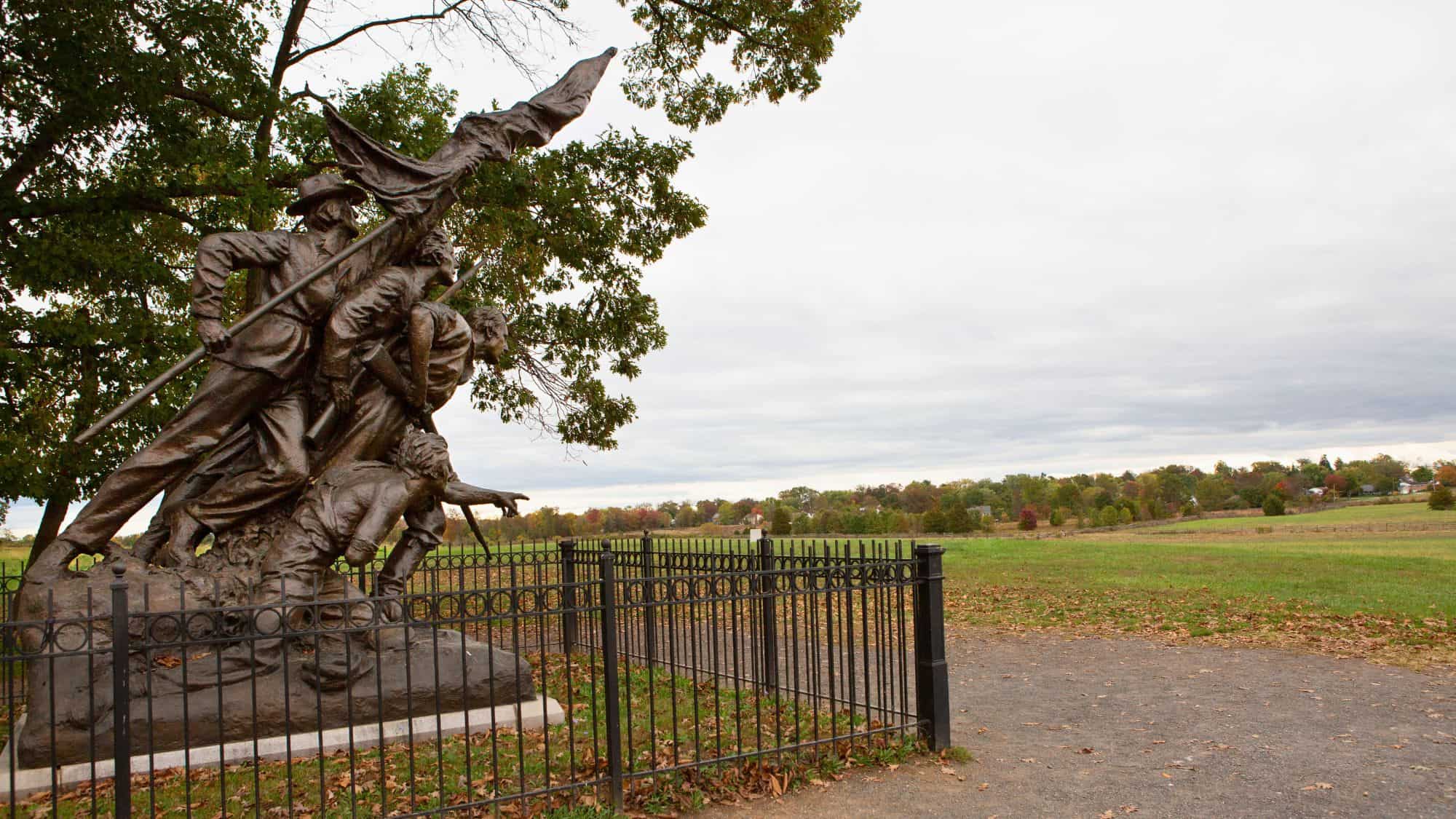 A dramatic bronze statue shows Confederate soldiers charging forward under a billowing flag, surrounded by a black metal fence and overlooking open fields.