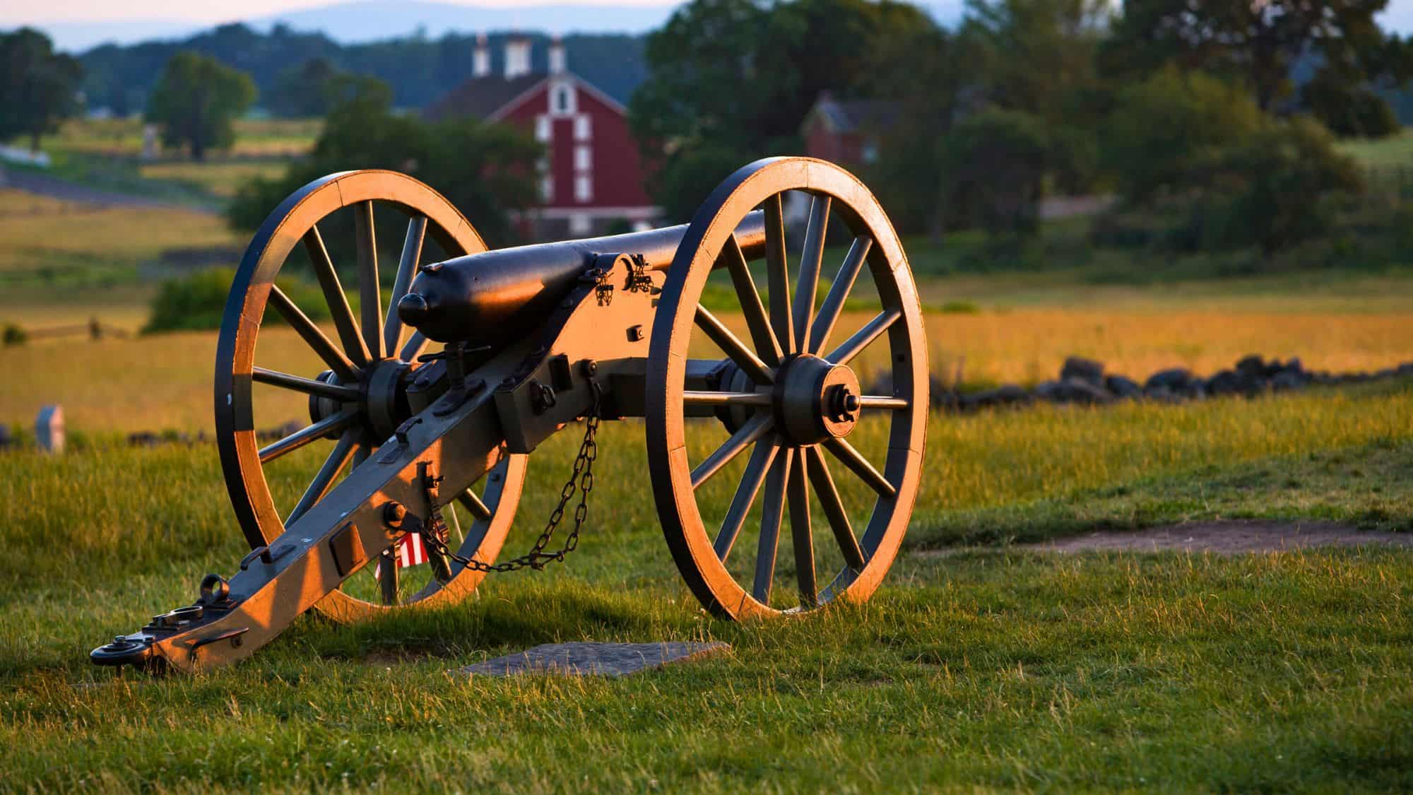 A Civil War-era cannon rests on a grassy field in the golden light of sunset, with a red barn in the background — marking the site of Pickett’s infamous charge.