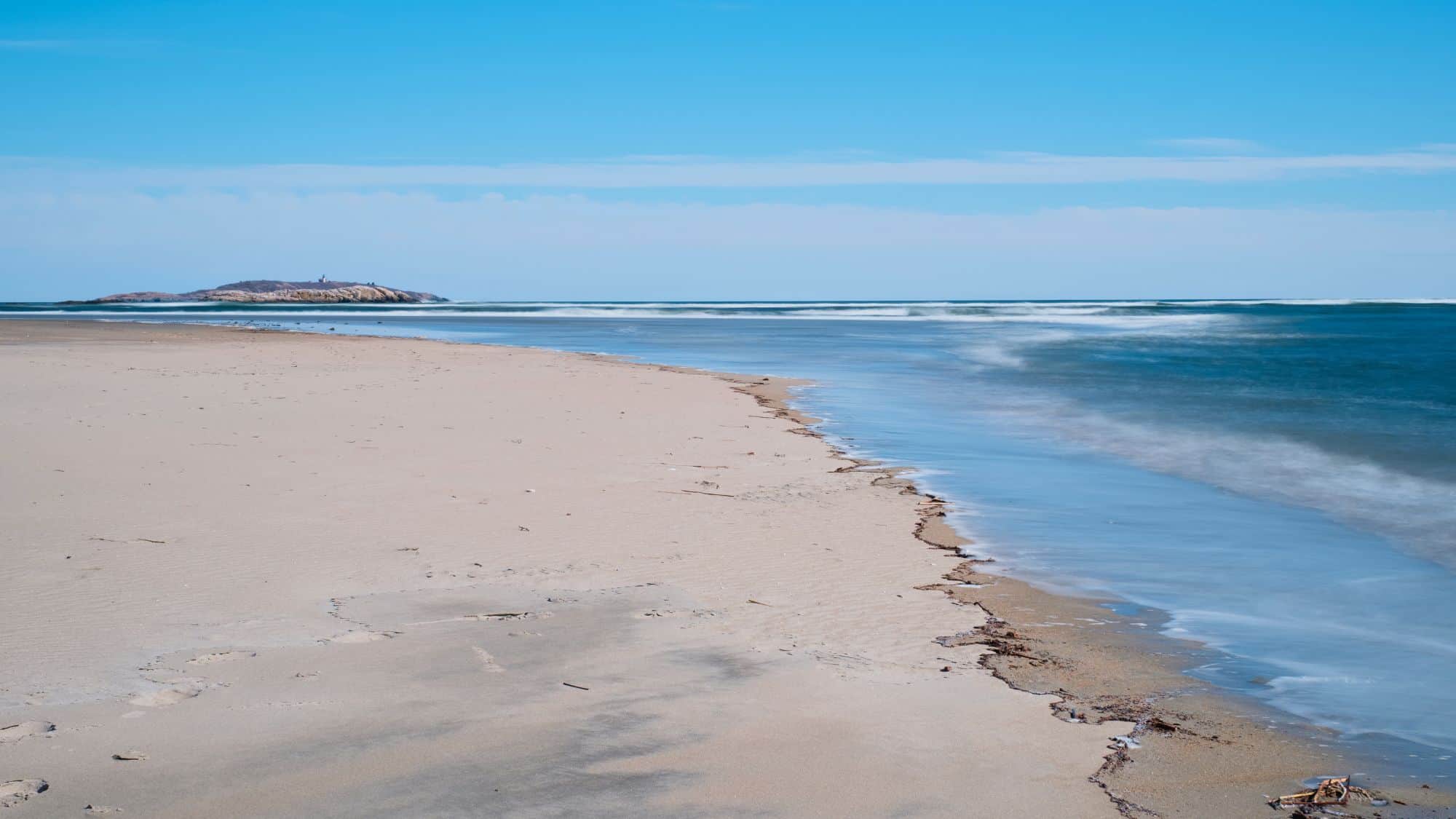 A narrow, sandy beach stretches along a calm, blue ocean with a small island visible in the distance under a bright sky.