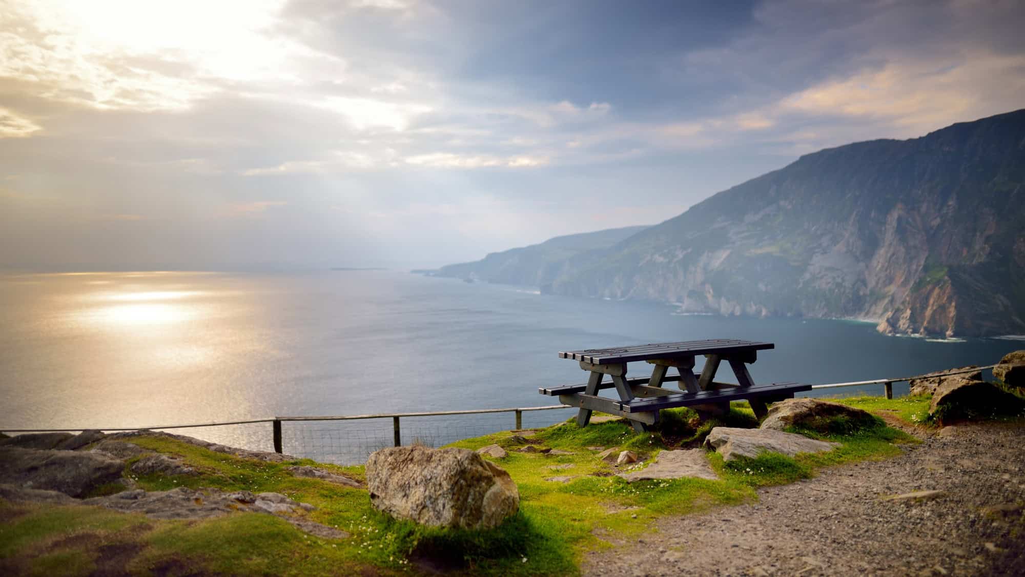 A wooden picnic table sits near the edge of the cliffs at Slieve League, offering a peaceful view of the Atlantic Ocean under soft, cloudy skies.

