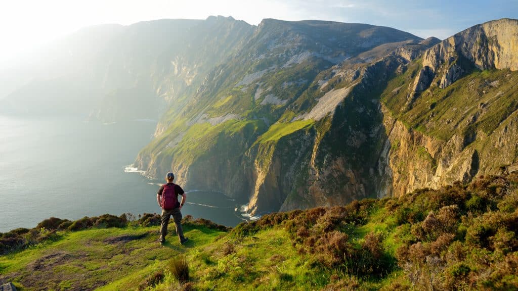 A solo hiker stands facing the vast expanse of Slieve League’s cliffs, lit by the golden glow of the setting sun with sheer cliffs and sea far below.