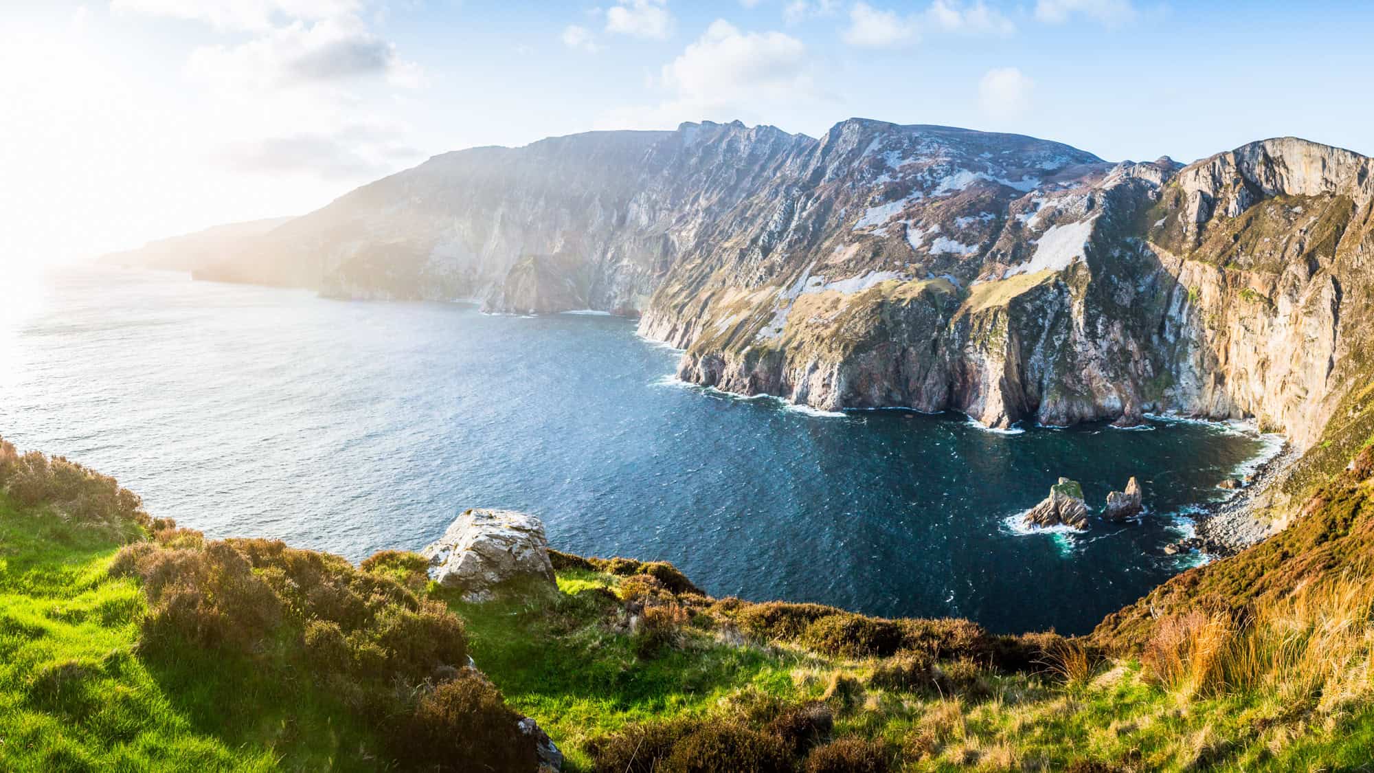Panoramic view of Slieve League Cliffs in Ireland at sunrise, where golden light spills over rugged sea cliffs meeting deep blue Atlantic waters.