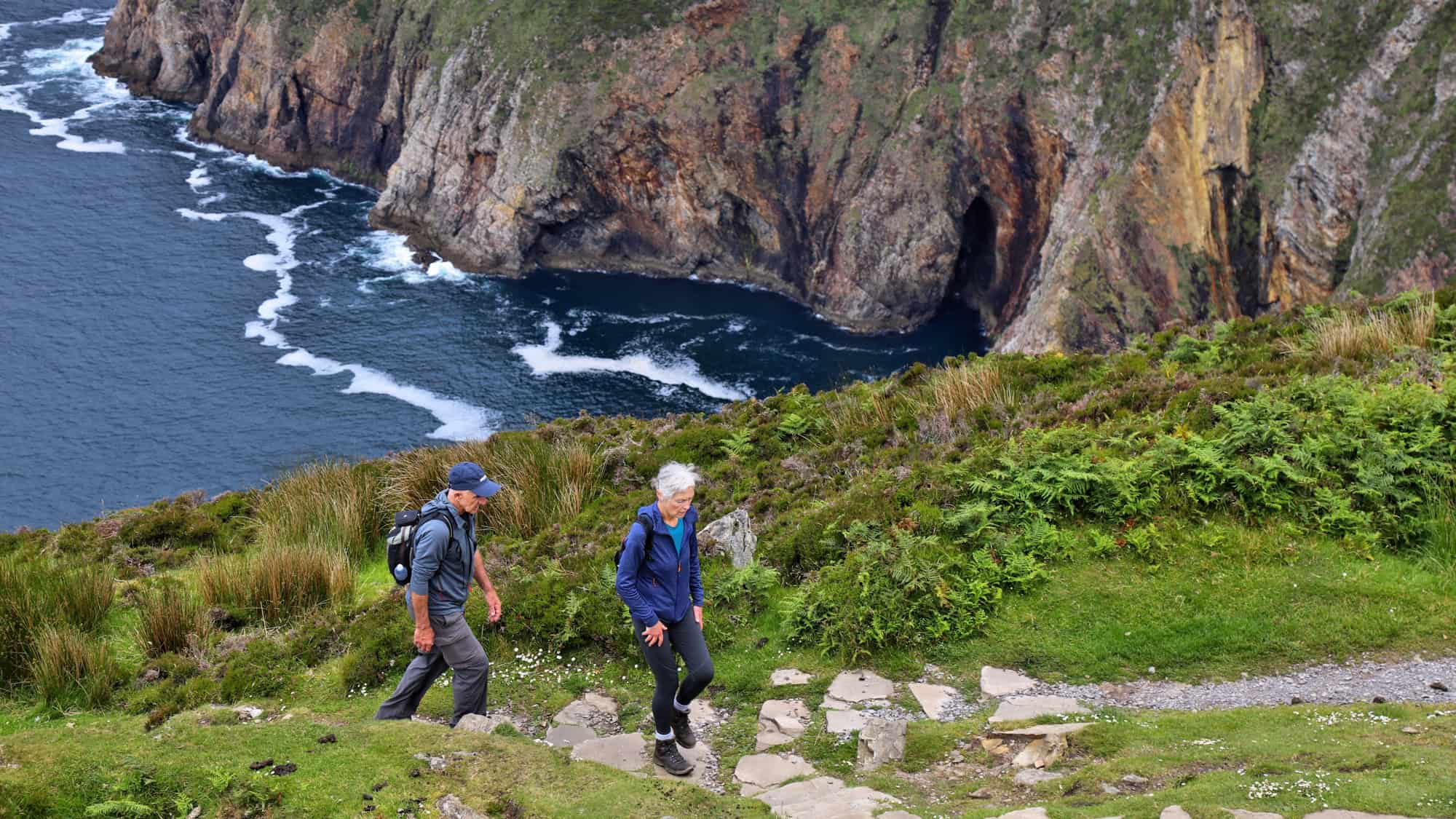Two hikers climb a grassy stone path overlooking the dramatic and rocky coastline of Slieve League Cliffs, with ocean waves crashing below.