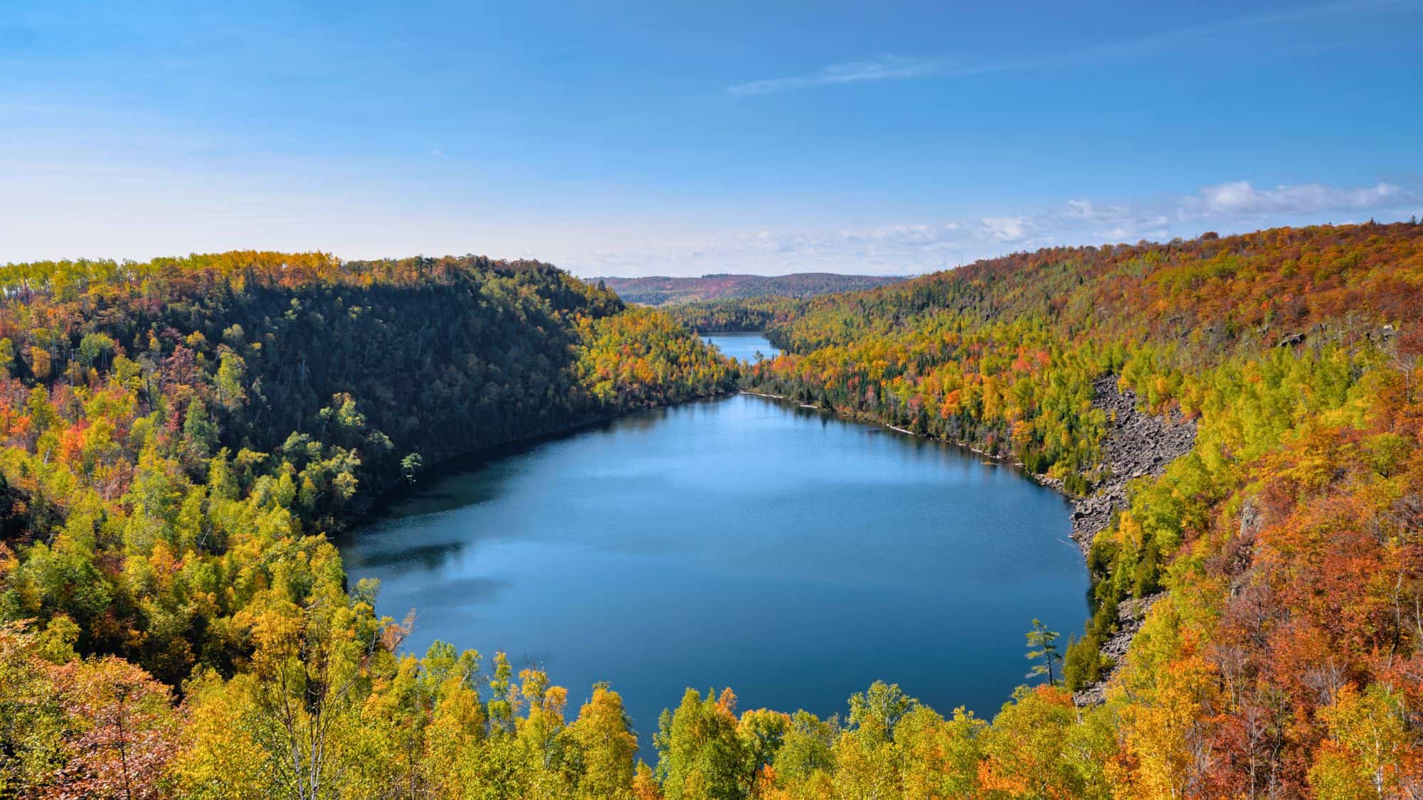 A vibrant forest in peak autumn color surrounds a calm, blue lake shaped like a heart, with rolling hills and distant waters extending to the horizon.