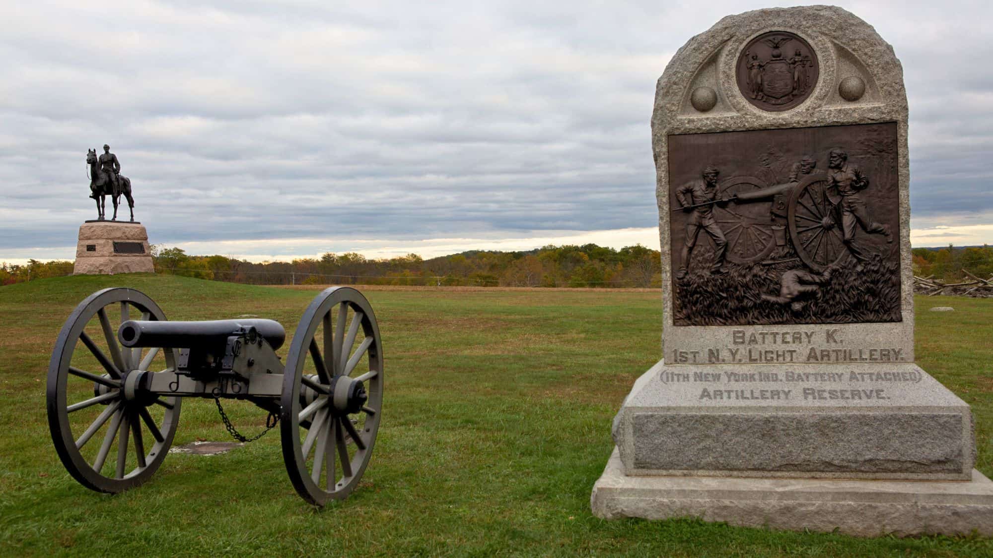 A cannon and a monument to Battery K, 1st N.Y. Light Artillery face the open field at The Angle, with a statue of a mounted officer visible in the distance on a hill.