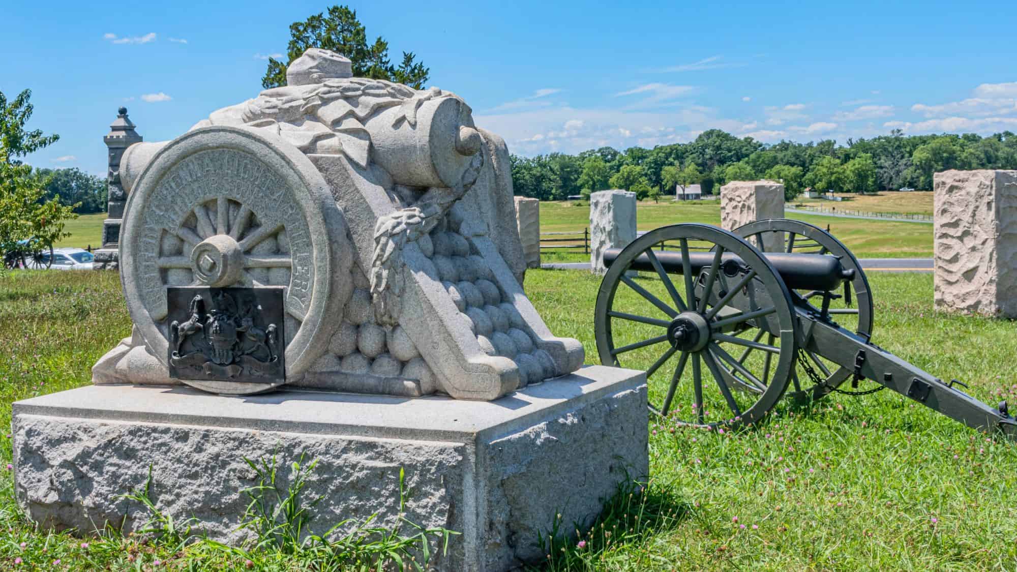 An elaborate cannon memorial sculpture made of stone sits beside an artillery piece on a grassy rise at the Peach Orchard battlefield sector under a clear summer sky.