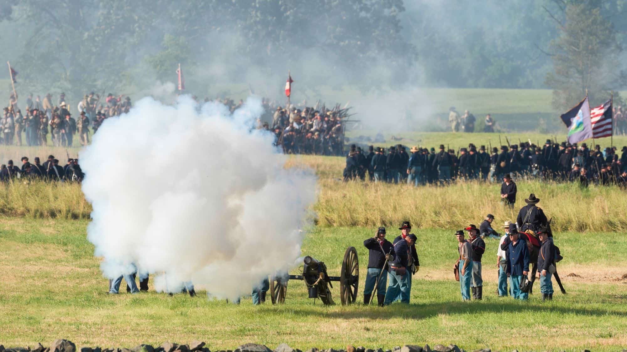 Civil War reenactors fire a cannon surrounded by smoke while opposing lines of infantry in blue and gray uniforms prepare for battle on the historic Wheatfield site.