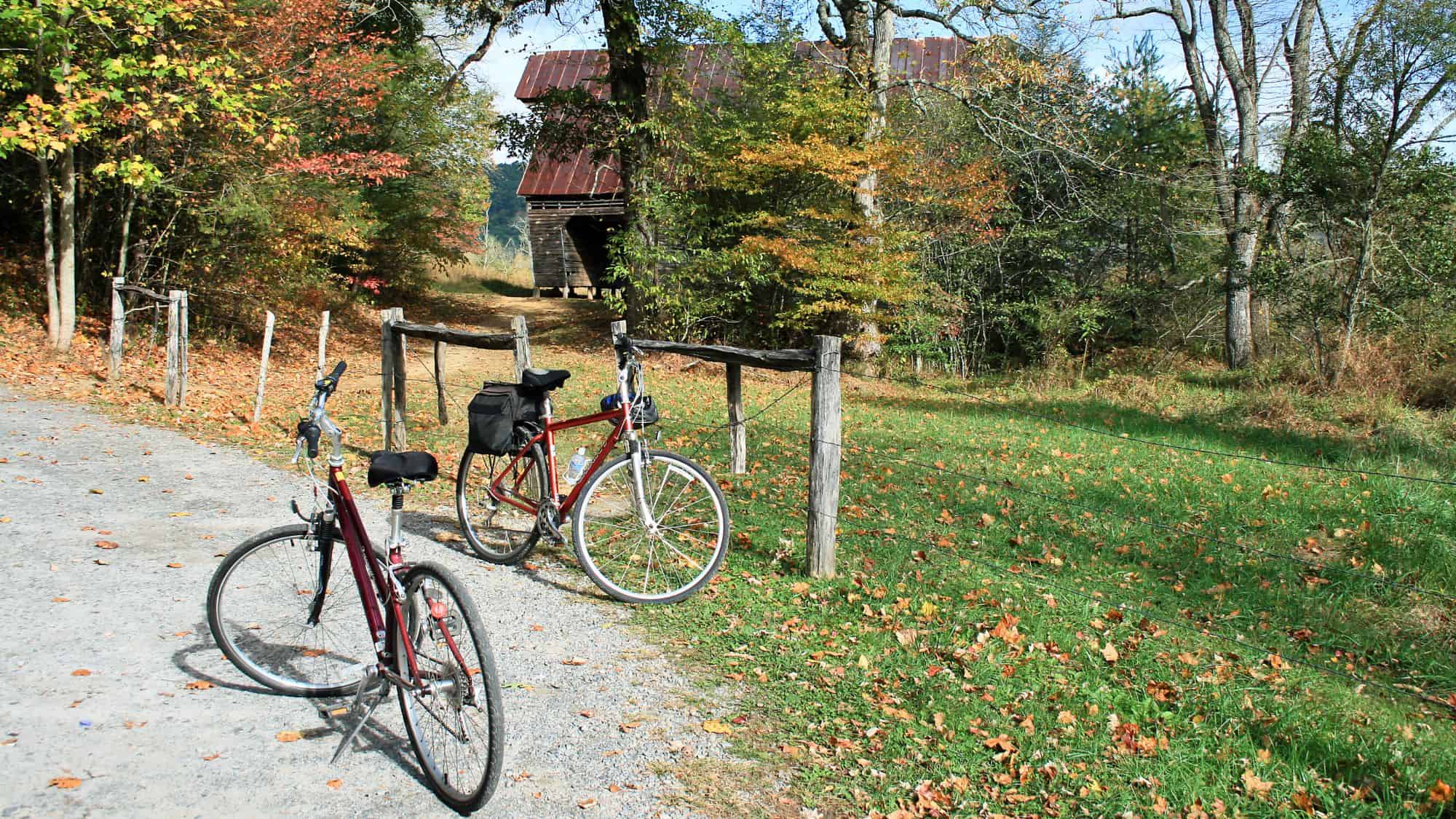 Two bicycles are parked along a rustic dirt path leading to an old barn, surrounded by trees in early autumn, suggesting a scenic pause on a countryside ride.