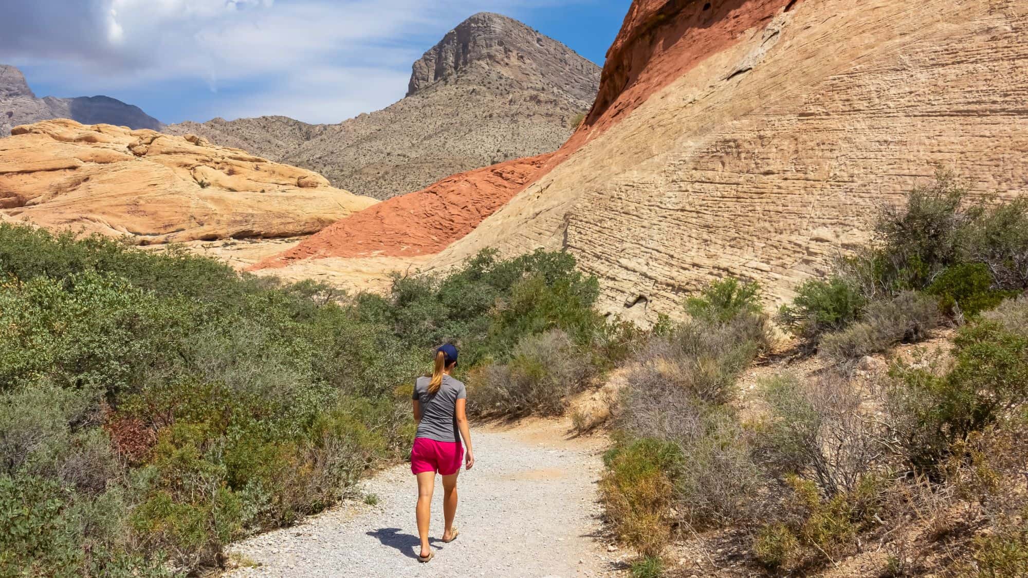 A hiker walks along a gravel trail surrounded by scrub brush and colorful sandstone formations in a sunlit desert mountain landscape.