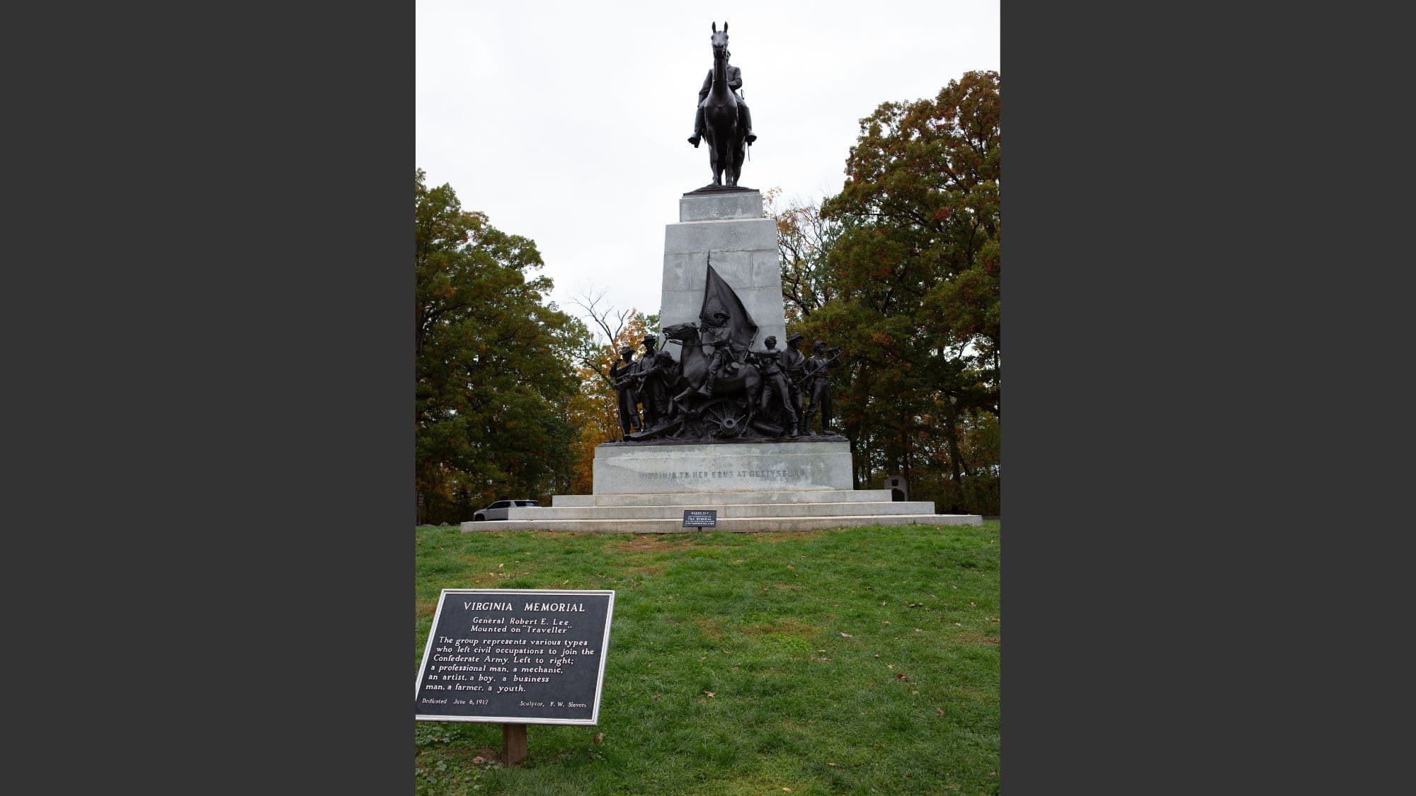 A towering monument features General Robert E. Lee on horseback atop a granite pedestal, with a bronze group sculpture below depicting a diverse group of men representing various professions who joined the Confederate army. A plaque in front explains the symbolism and notes the memorial’s dedication date of June 8, 1917.