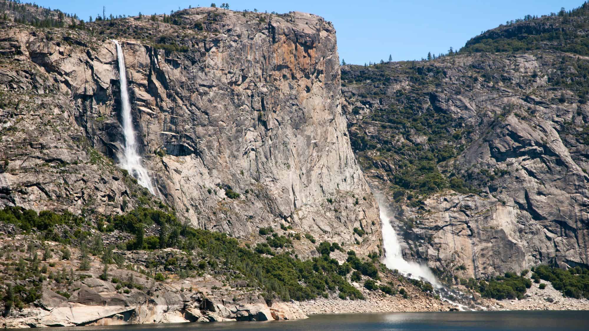 Twin waterfalls cascade down sheer granite cliffs into the Hetch Hetchy Reservoir below, surrounded by lush foliage and rocky outcrops.