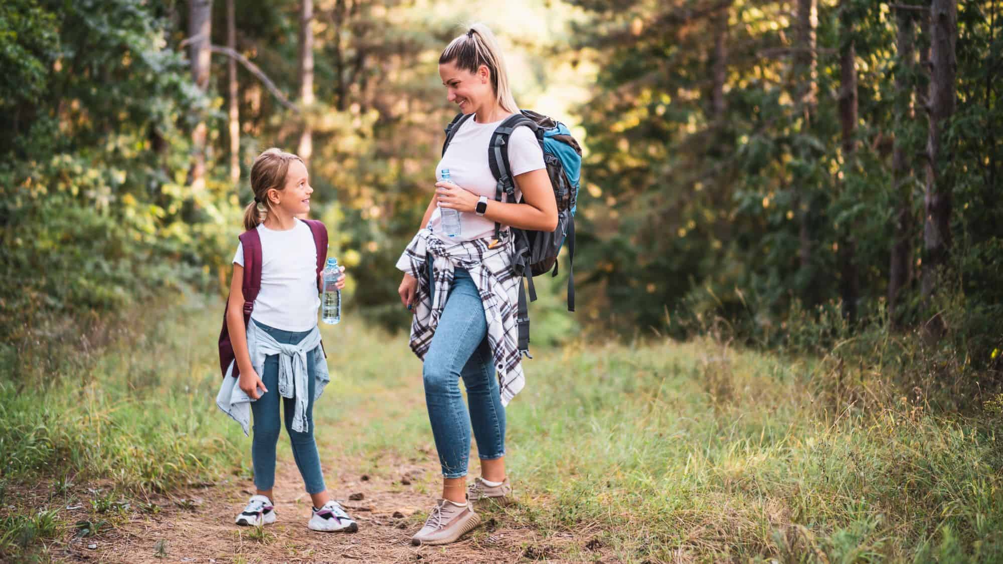 Mother and daughter on a forest trail taking a break from hiking, both carrying backpacks and holding water bottles while smiling at each other.